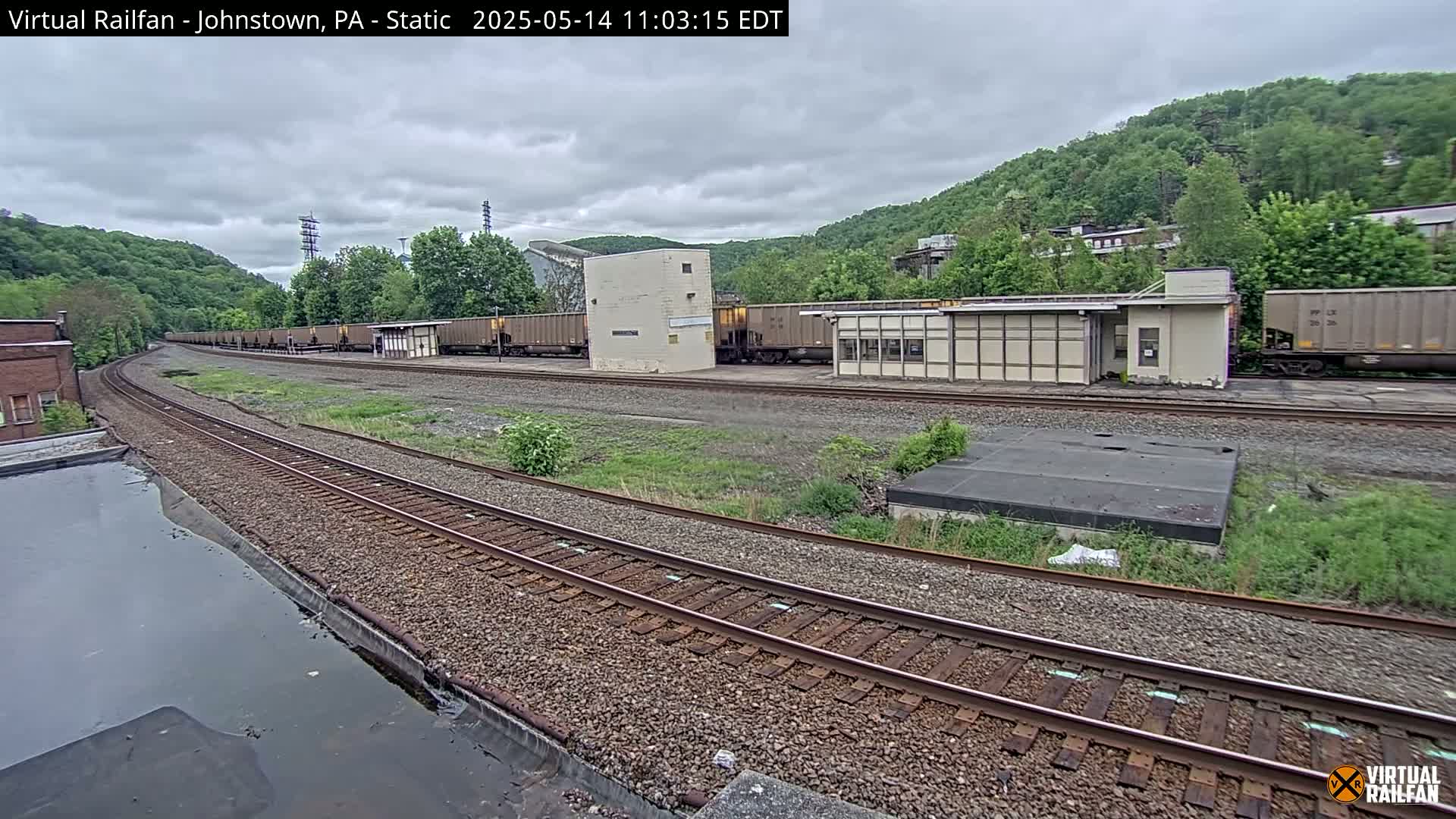 A long line of freight cars sits on a curving railway track next to a small building, under an overcast sky, against a backdrop of lush green hills.