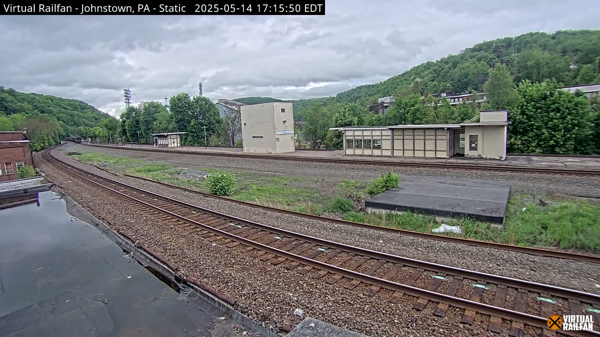 Train tracks curve gently through a train yard under an overcast sky, with a small building and overgrown vegetation visible nearby.