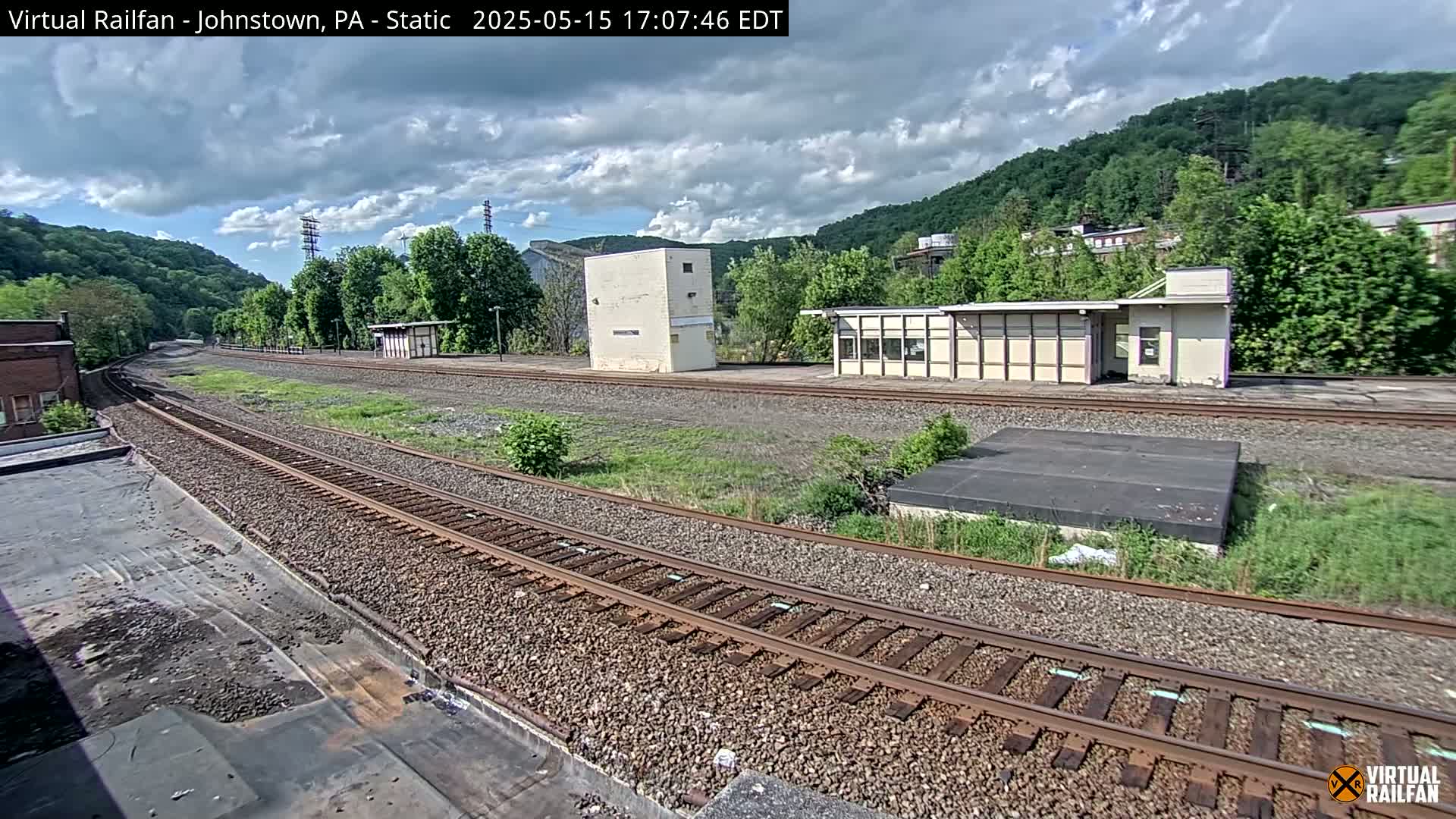 A partly cloudy sky overlooks railroad tracks that curve gently to the left, passing by several small buildings and a hill covered in lush green trees.