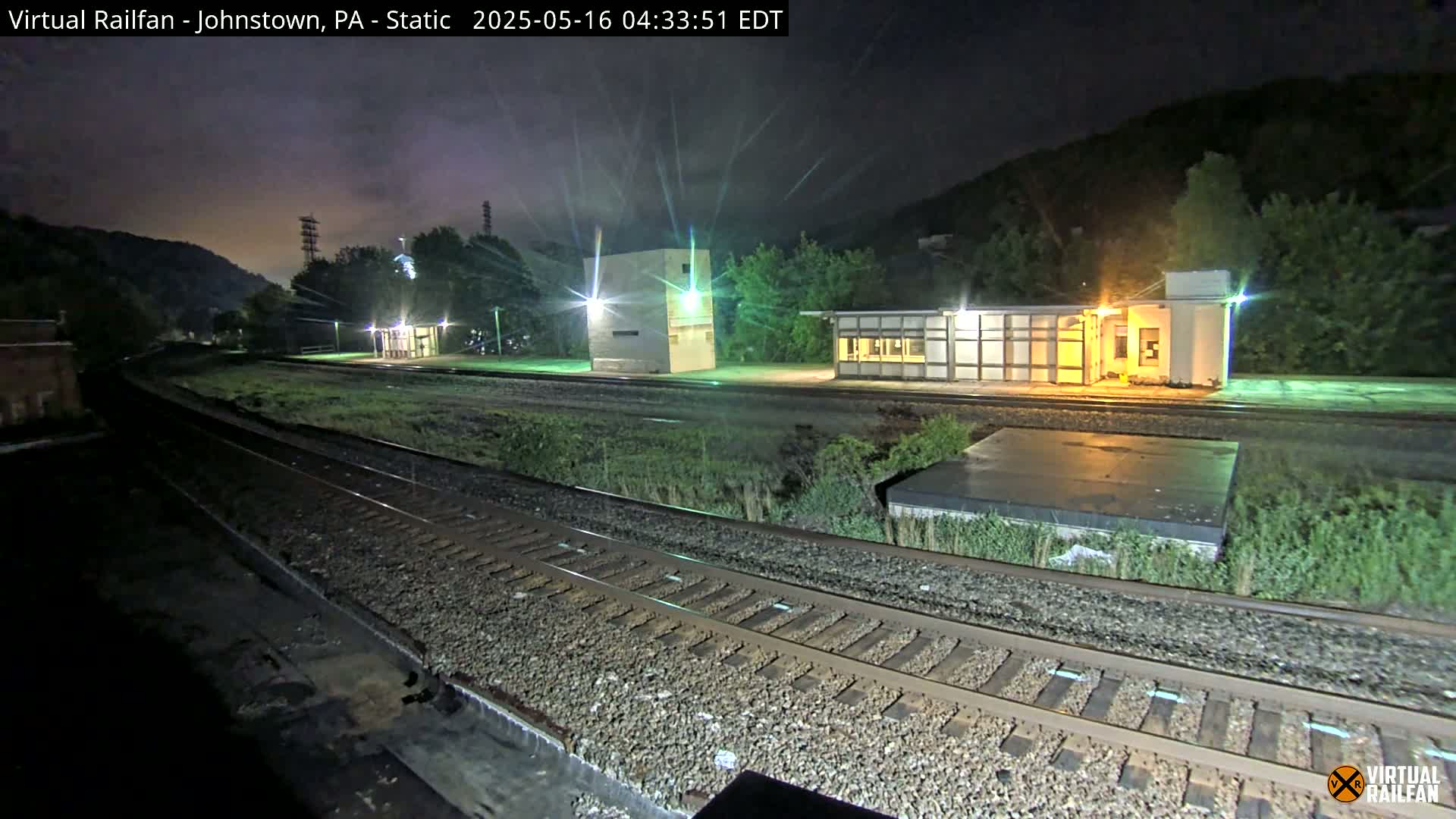 A nighttime view of a train station platform with multiple buildings and railway tracks, under a dark, cloudy sky.