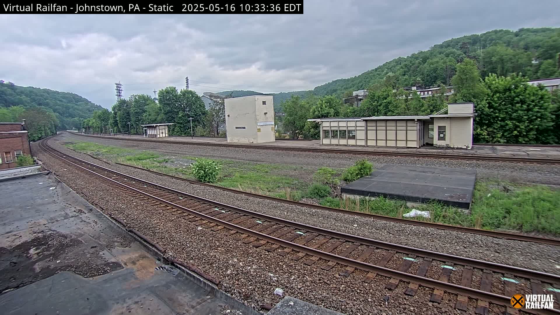 A view of train tracks curving through a valley on an overcast day, with several small buildings and overgrown vegetation alongside the tracks.