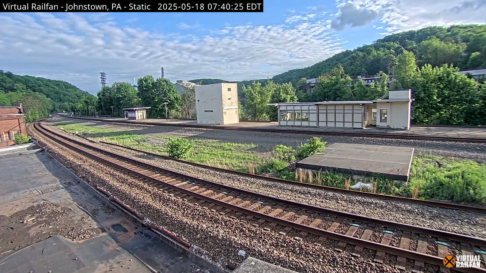 A partly cloudy sky overlooks a train station with two small buildings, railroad tracks curving away from the viewer, and lush green hills in the background.