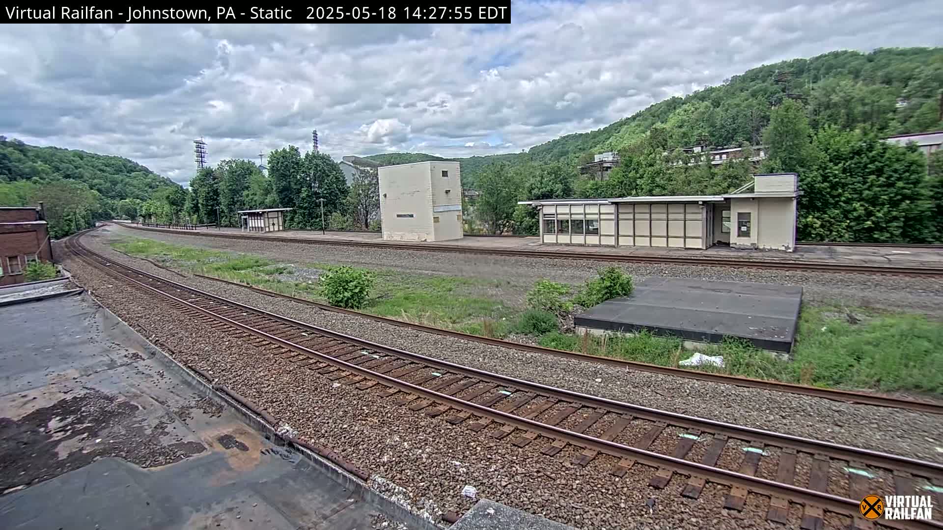 A train track curves through a partly cloudy landscape, passing by several small buildings and a hillside covered in lush green trees.