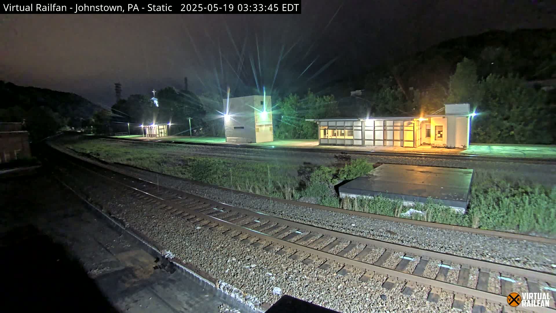 A nighttime view of a deserted train station platform with train tracks, and several small buildings under a dark sky.