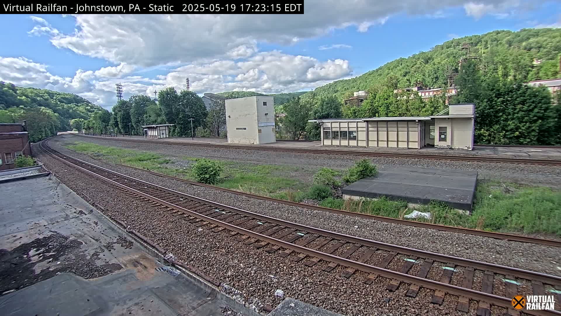 A partly cloudy day shows a train track curving through a grassy area with a few small buildings and hills covered in lush green trees in the background.