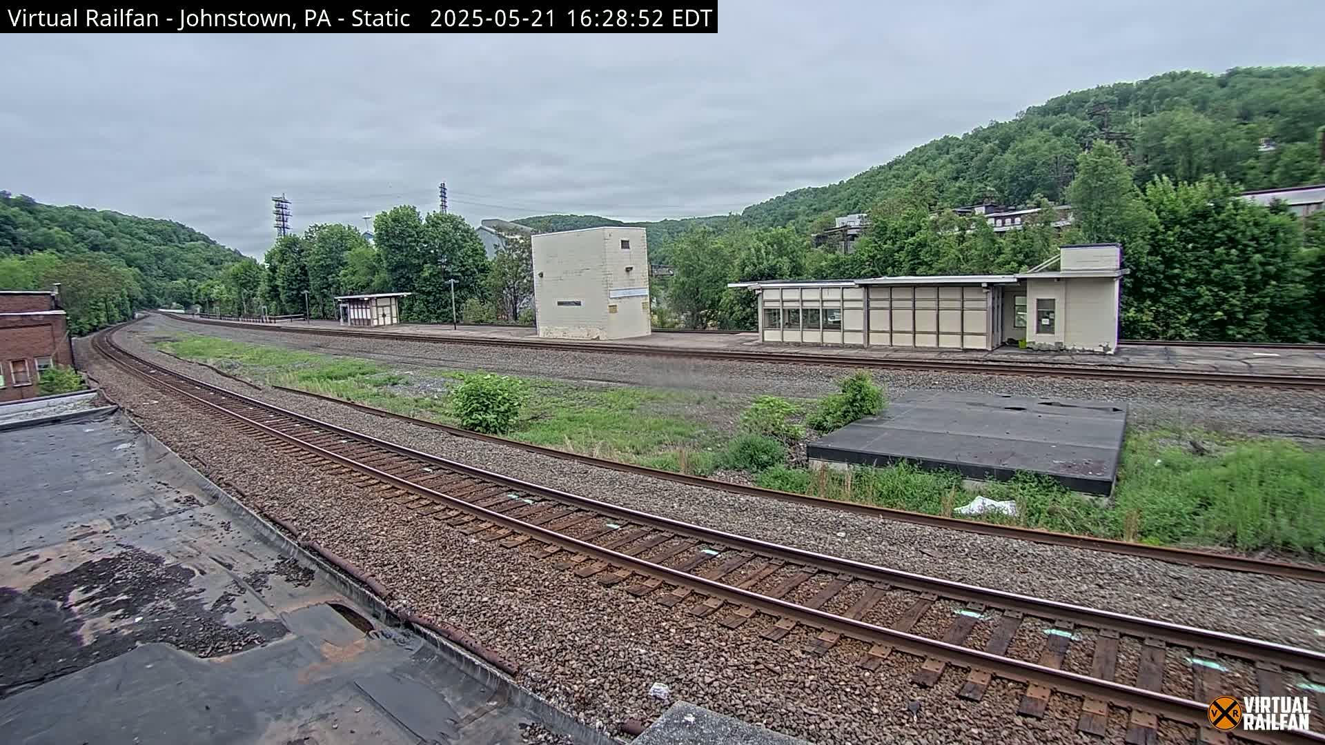 A train track curves gently through a grassy area with small buildings and overgrown vegetation, all set against a backdrop of verdant hills under an overcast sky.