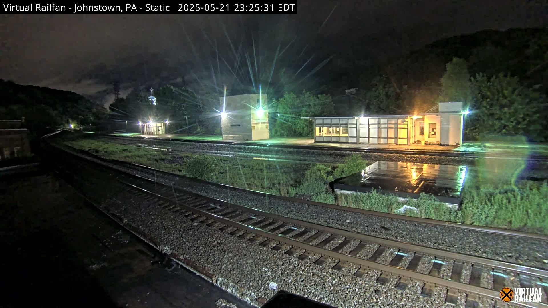 A nighttime view of a rain-slicked train station platform and tracks, with several small buildings illuminated under a dark, cloudy sky.