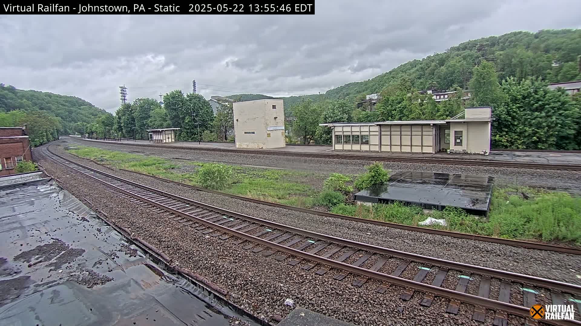 A railroad track curves through a verdant valley under an overcast sky, passing alongside several small, light-colored structures.