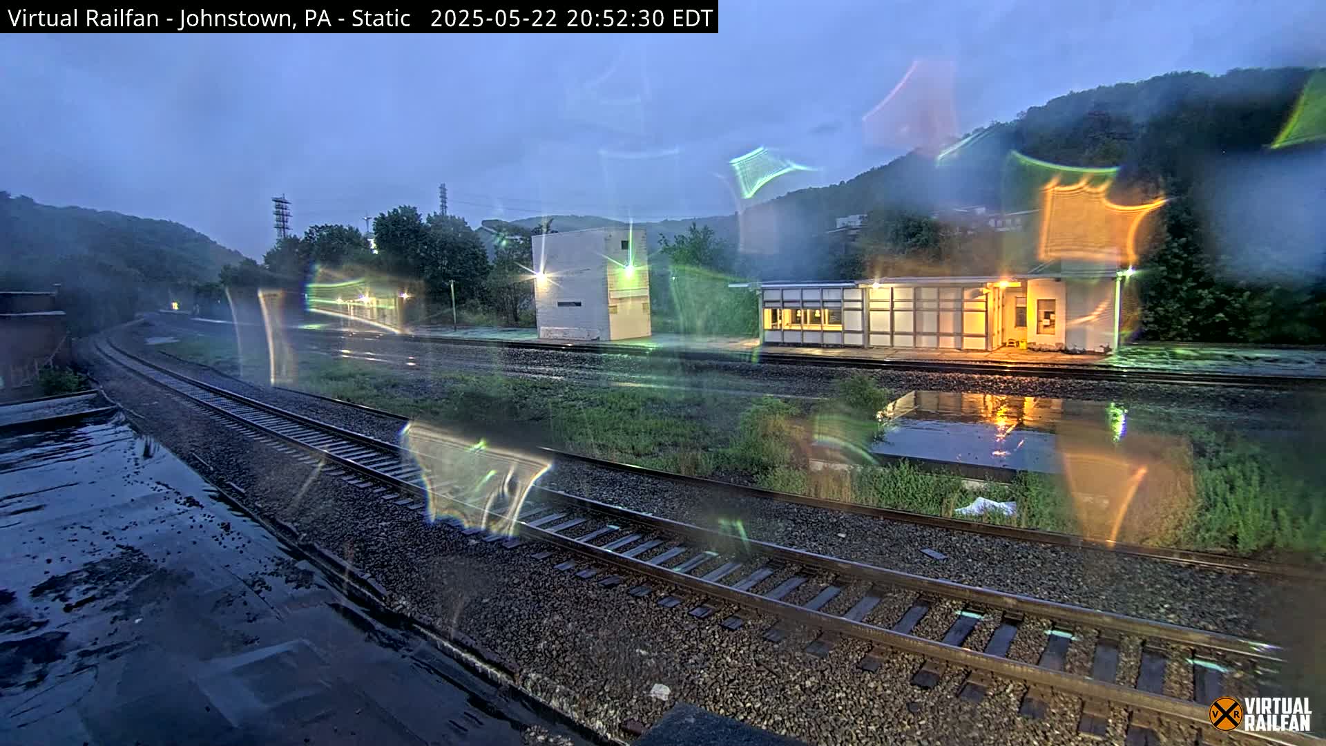 A rain-slicked train station and railway tracks are visible at twilight, with hills in the background.