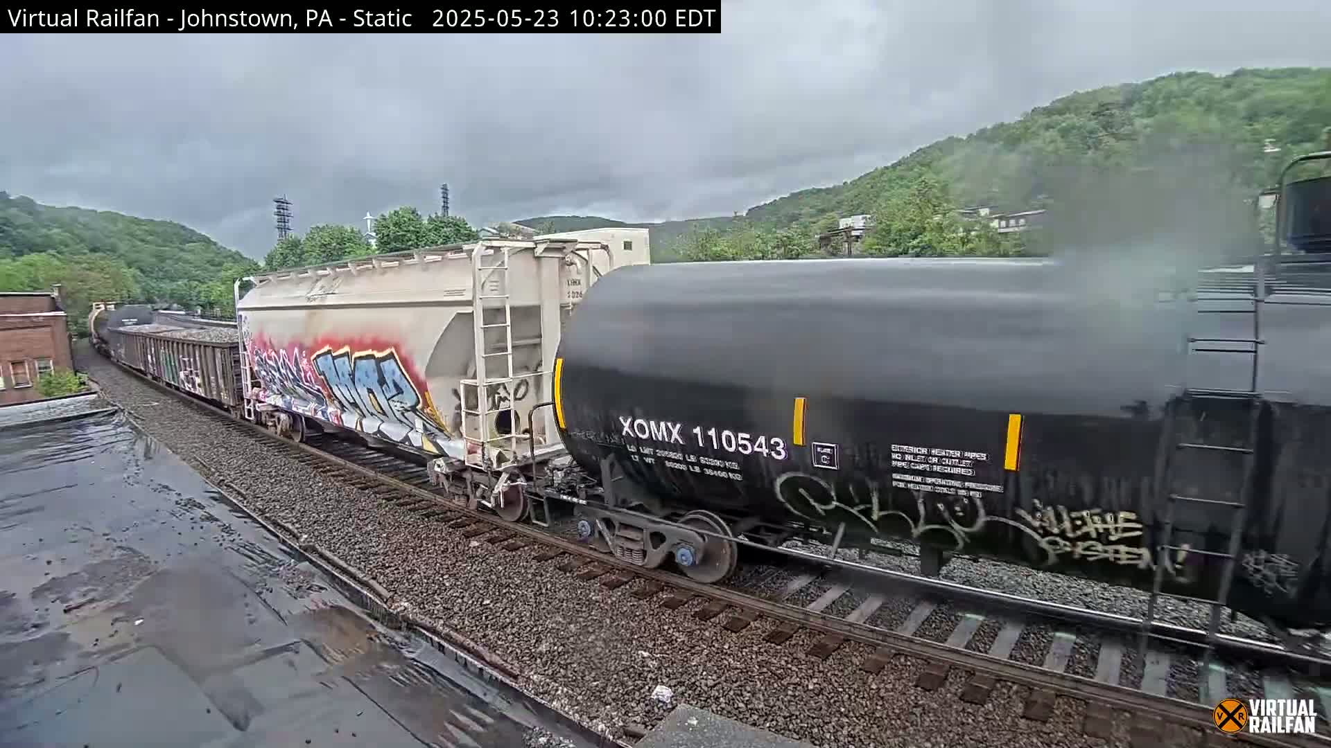 A long freight train with graffiti-covered cars passes by on a rainy day, with hills visible in the background.