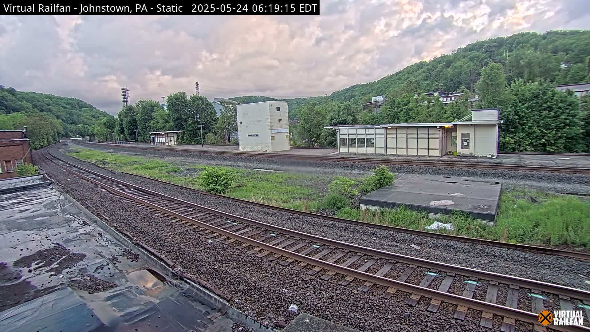 A view of train tracks curving away from the camera, past several small buildings and overgrown areas, under a partly cloudy sky.