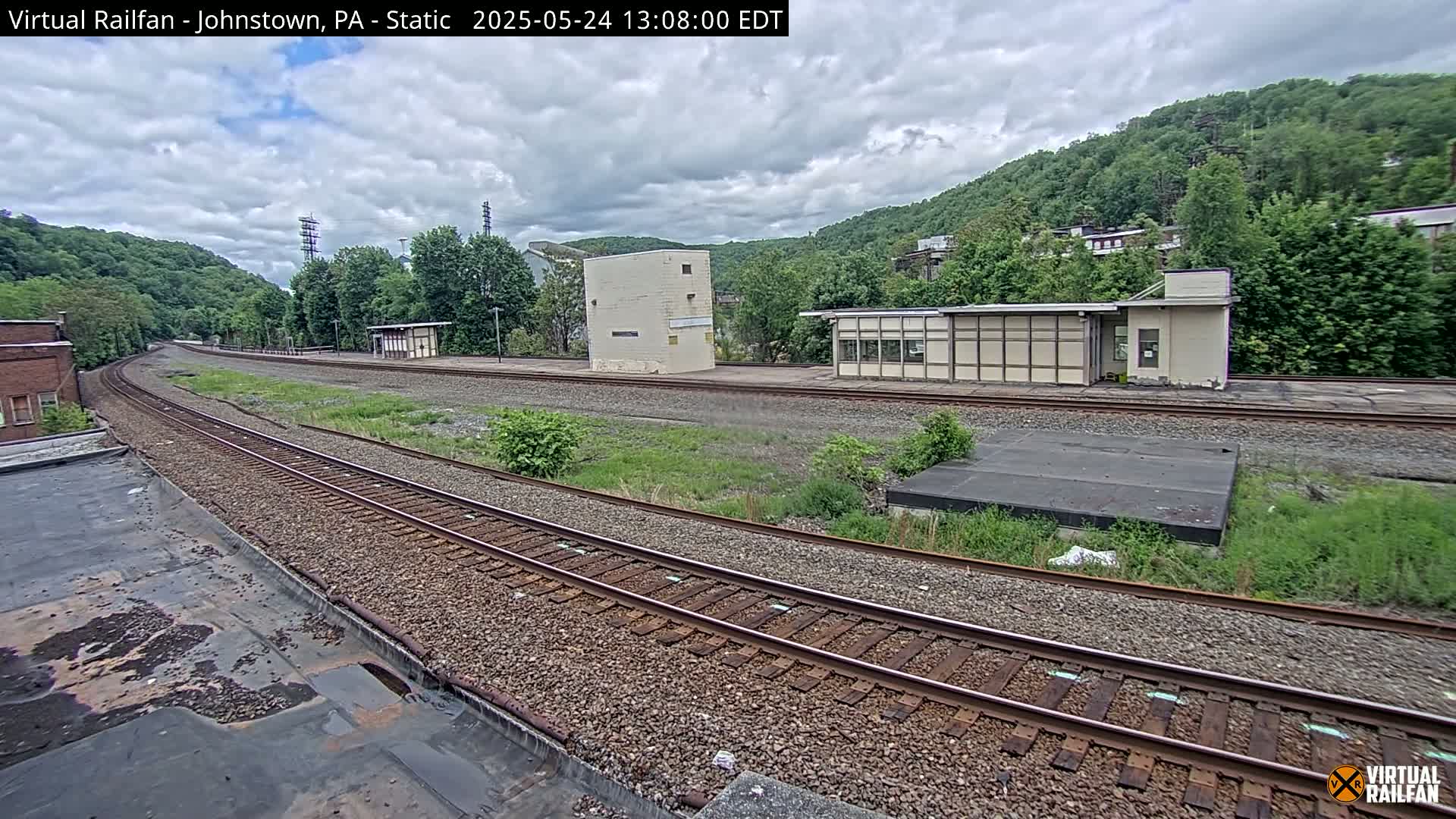 A curving railway track stretches into the distance, flanked by overgrown vegetation, small buildings, and a hillside covered in lush green trees under a partly cloudy sky.