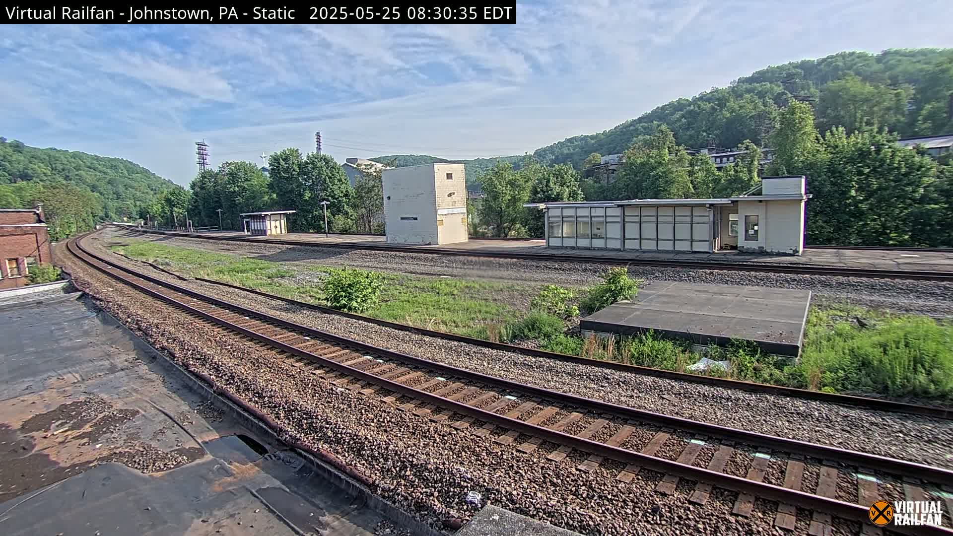 A train station with two small buildings sits beside curving train tracks that extend into a lush, green hillside under a partly cloudy sky.