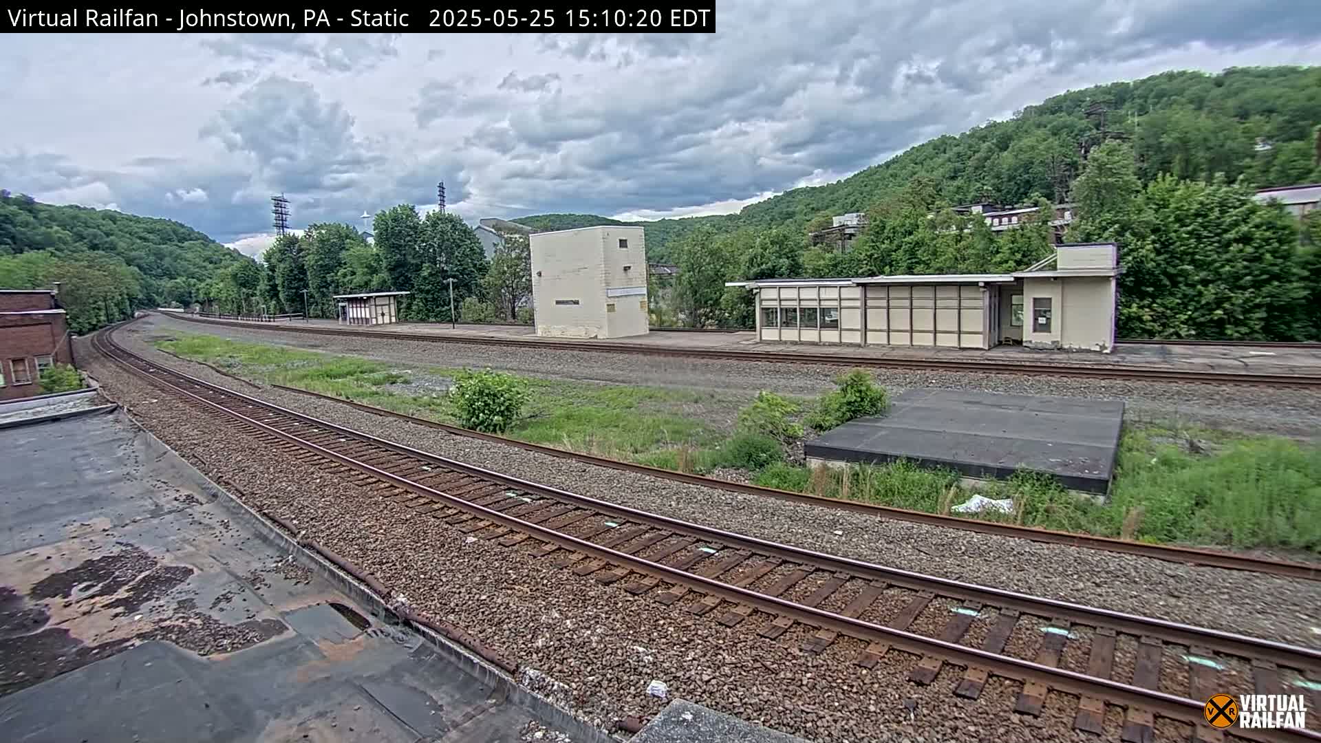 A curving railroad track winds through a grassy area past several small buildings, with a hillside covered in lush green trees under an overcast sky.