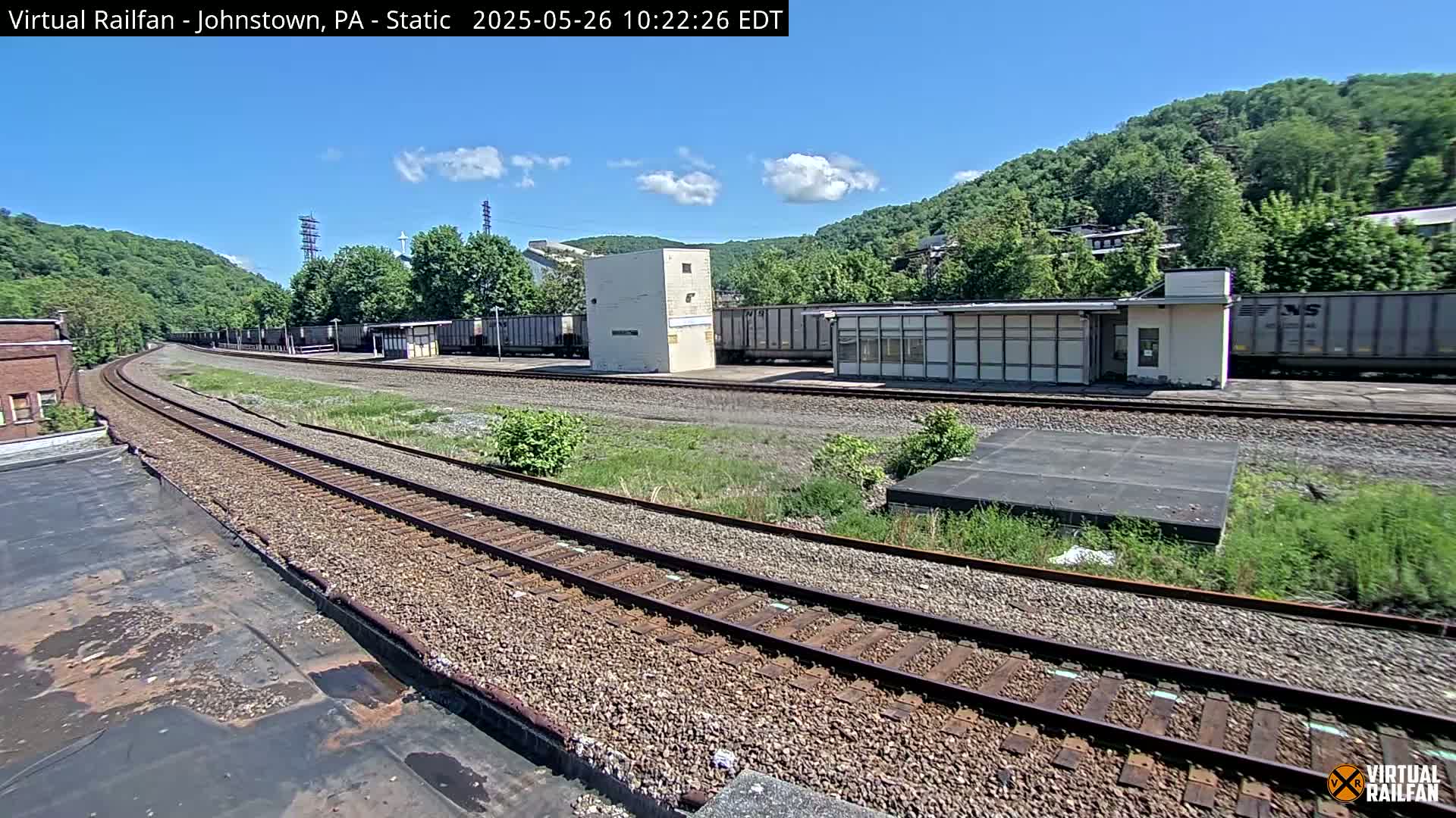 A sunny day reveals a train yard with several railcars, a small building, and a curving railway track winding through a hilly, green landscape.