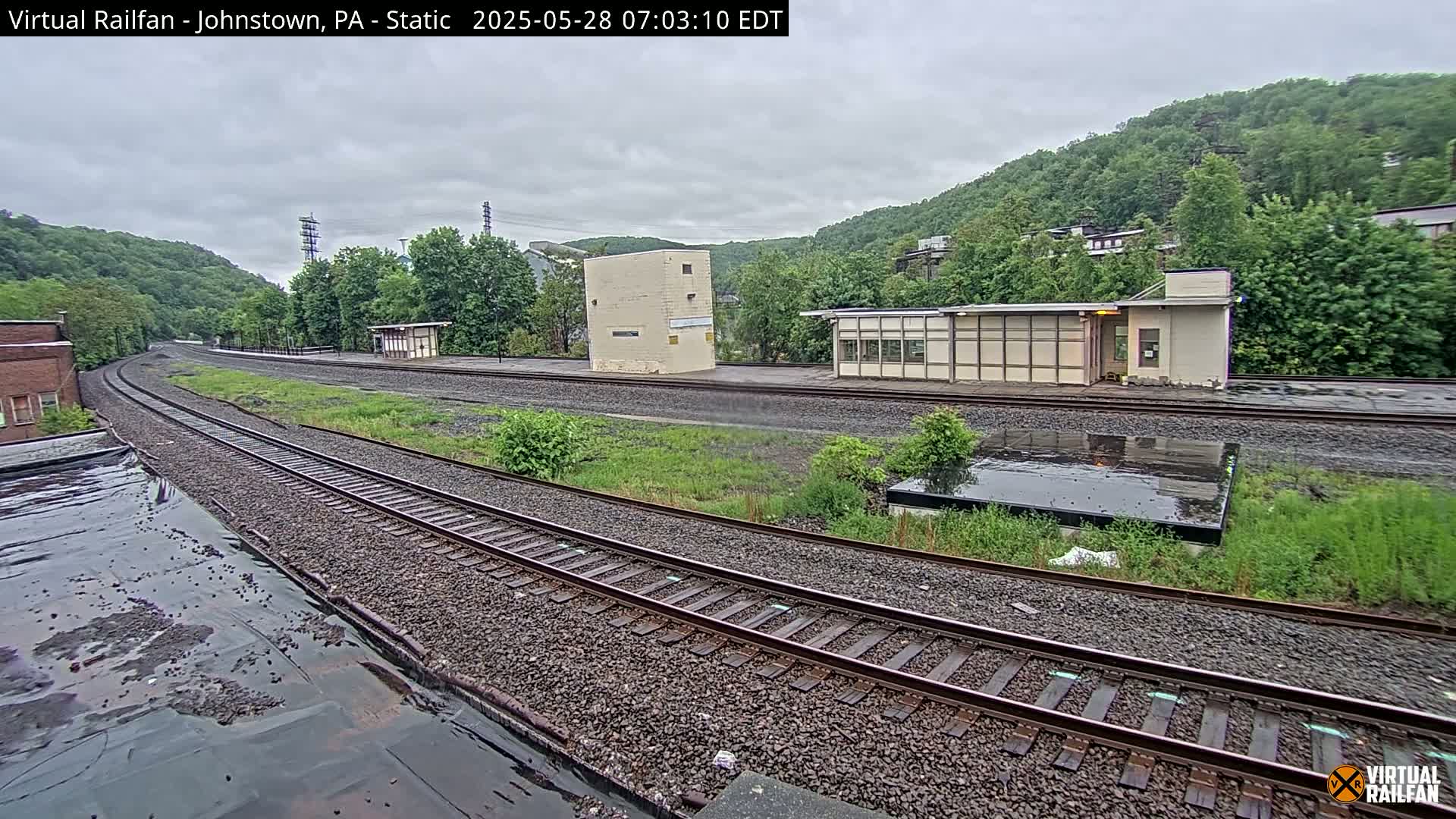A wet train station with multiple tracks curves gently around a small building and several overgrown areas, set against a backdrop of lush green hills under an overcast sky.
