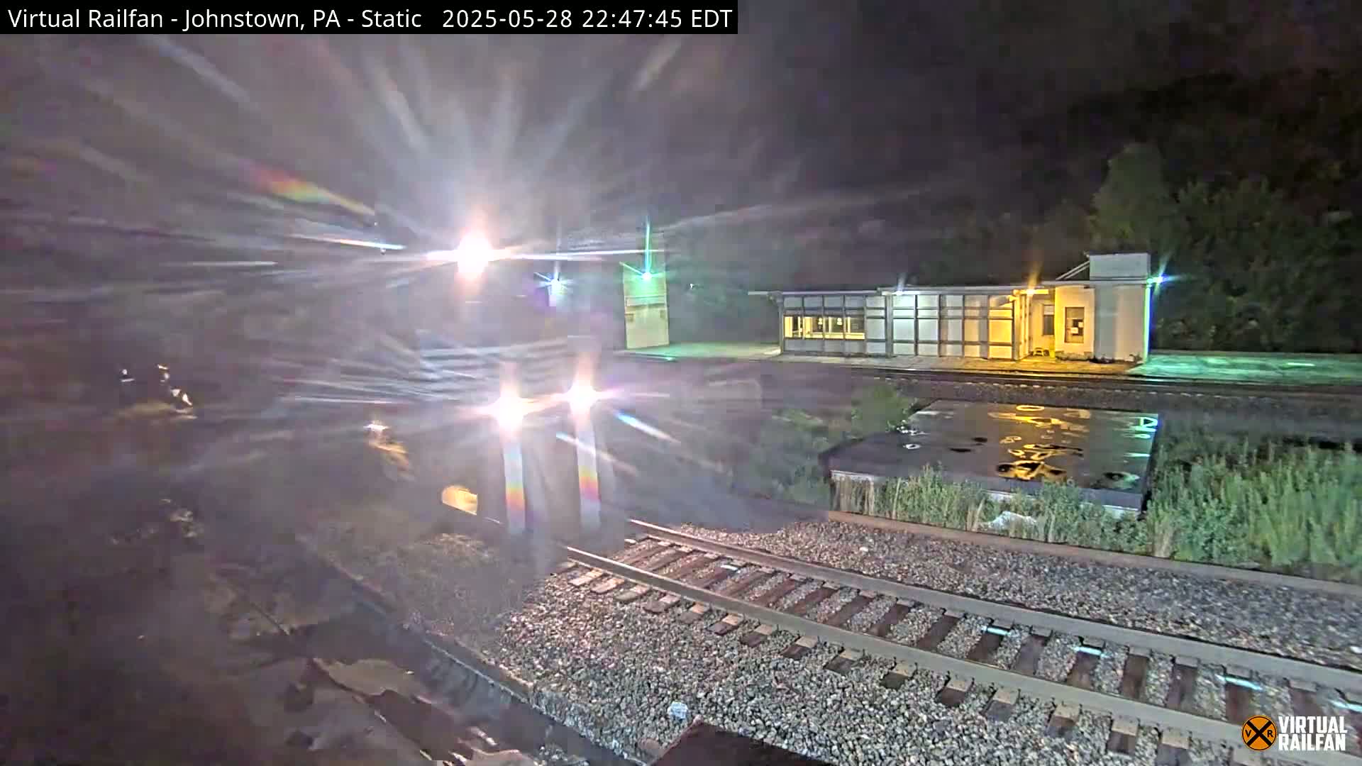 A nighttime view of a train approaching a station platform, with some wet areas visible near the tracks.