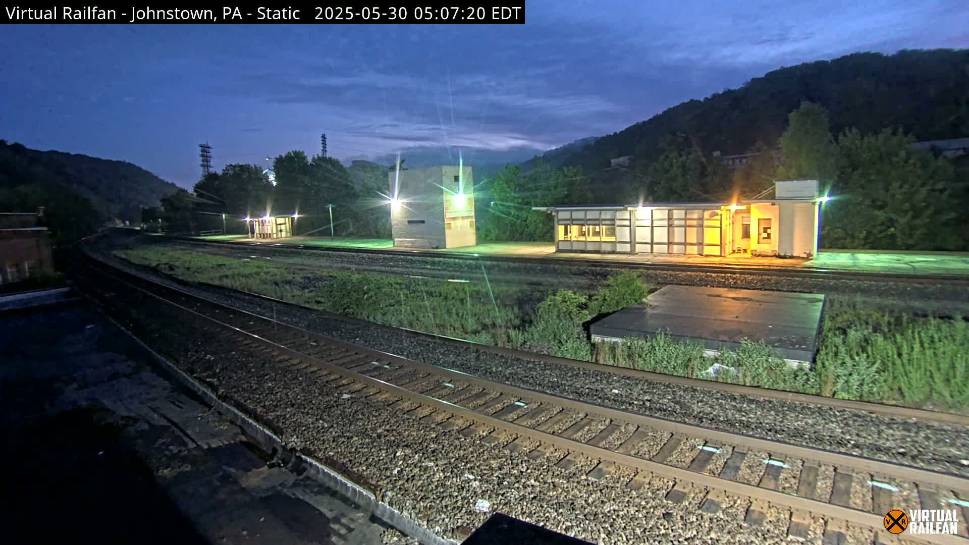 A nighttime view of a train station with several structures, railroad tracks, and hills in the background under a twilight sky.