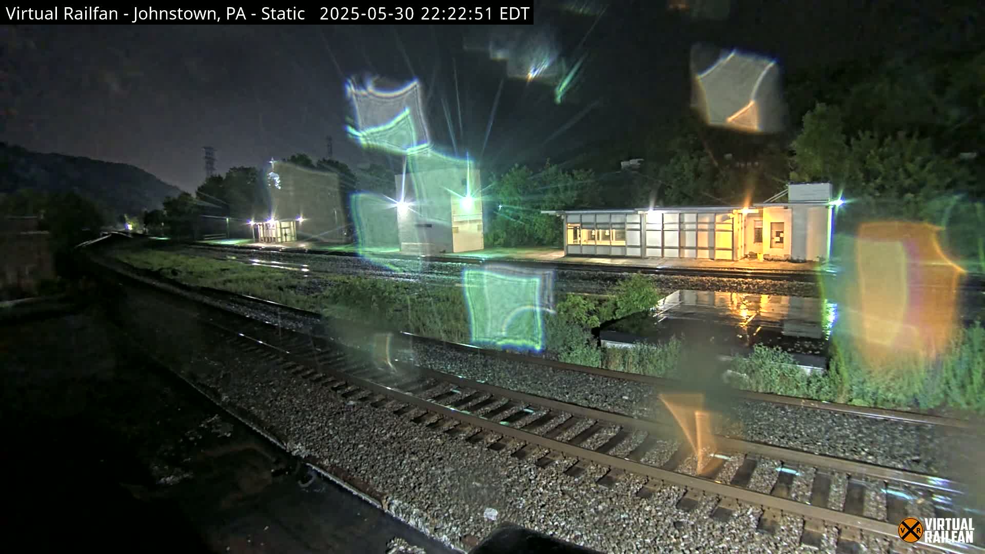 A nighttime view of a rain-slicked train platform and tracks, with a small building and surrounding vegetation visible under illuminated conditions.