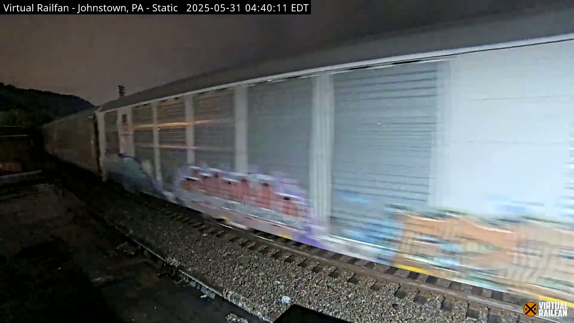 A long freight train with graffiti on some cars moves quickly along train tracks at night.