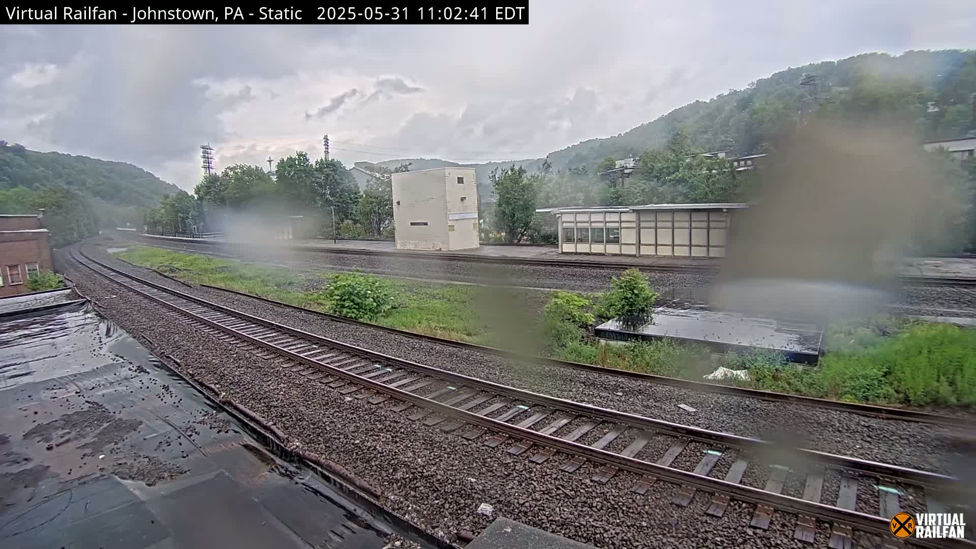 A rainy view of a train station with multiple train tracks, surrounded by green hills and some buildings.