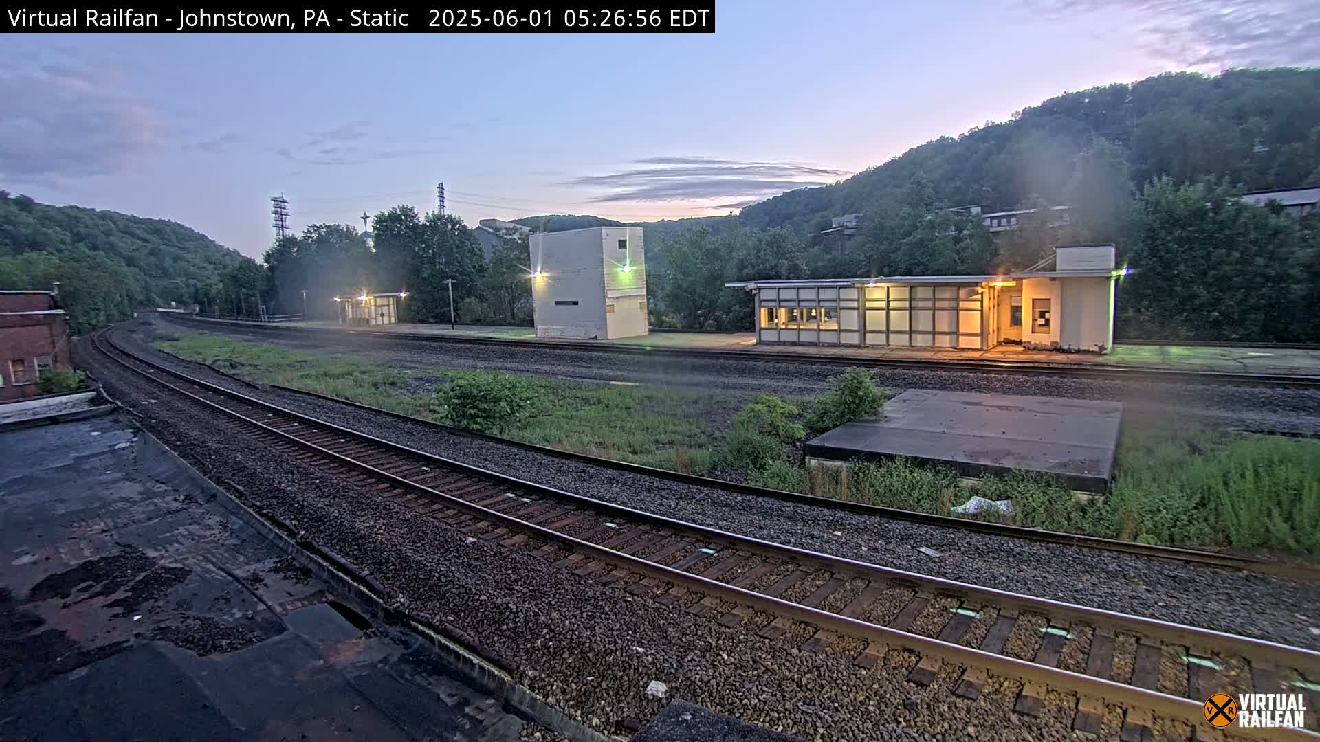 A twilight view of a train station with two buildings alongside curving train tracks, set against a backdrop of lush green hills.
