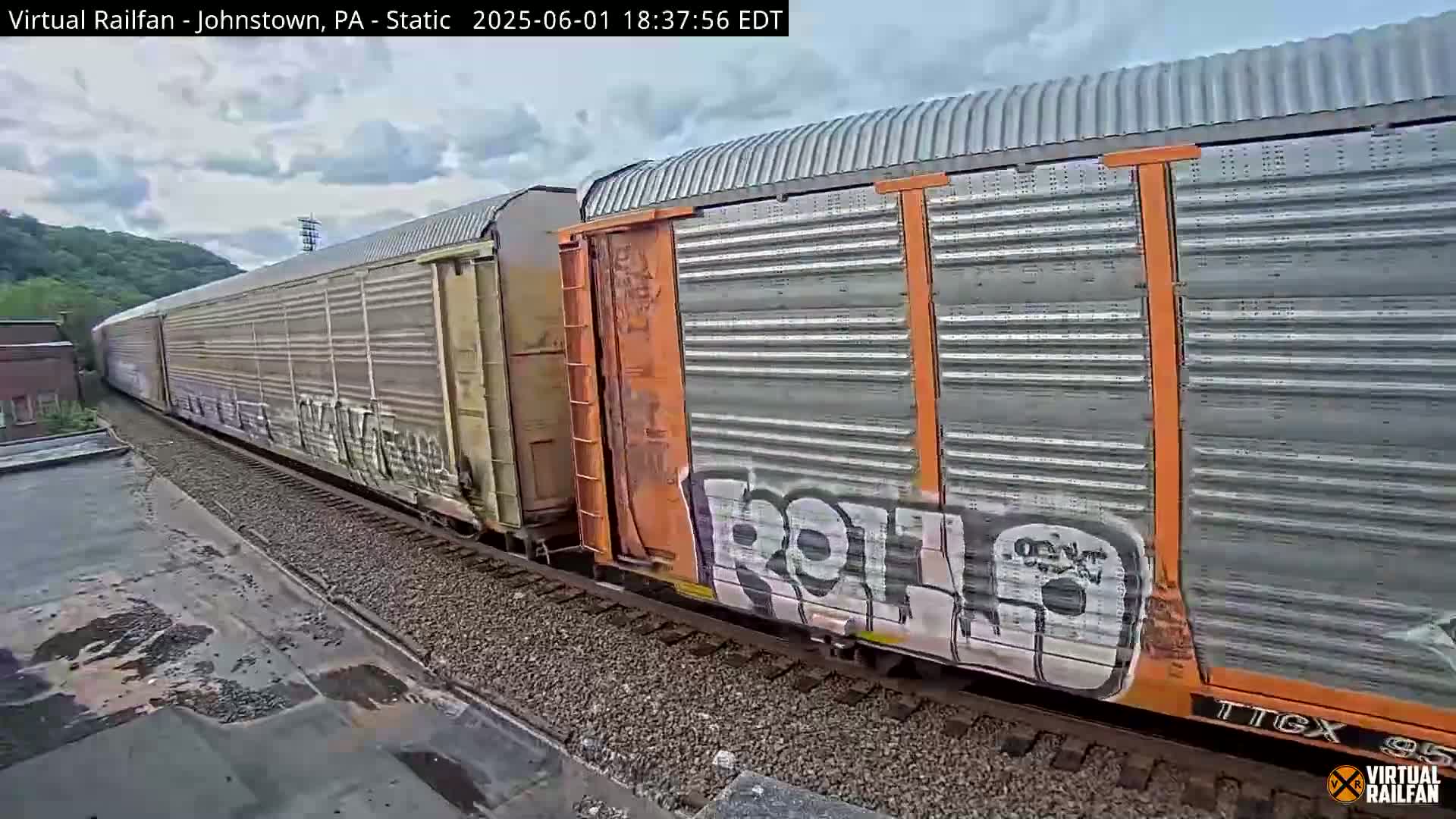 A long freight train with graffiti on some of its cars moves along railroad tracks under a partly cloudy sky.