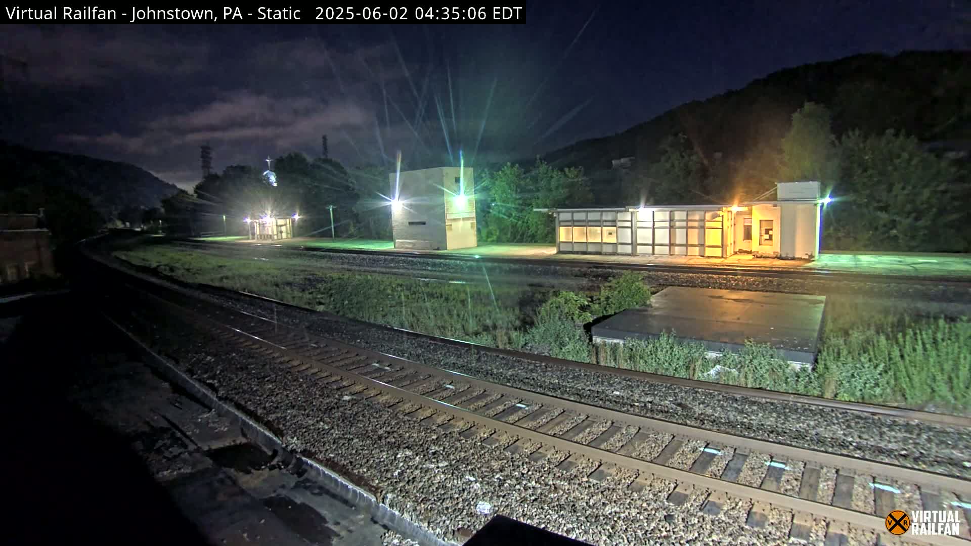 A nighttime view of a train station with several buildings illuminated, alongside train tracks, under a partly cloudy sky.