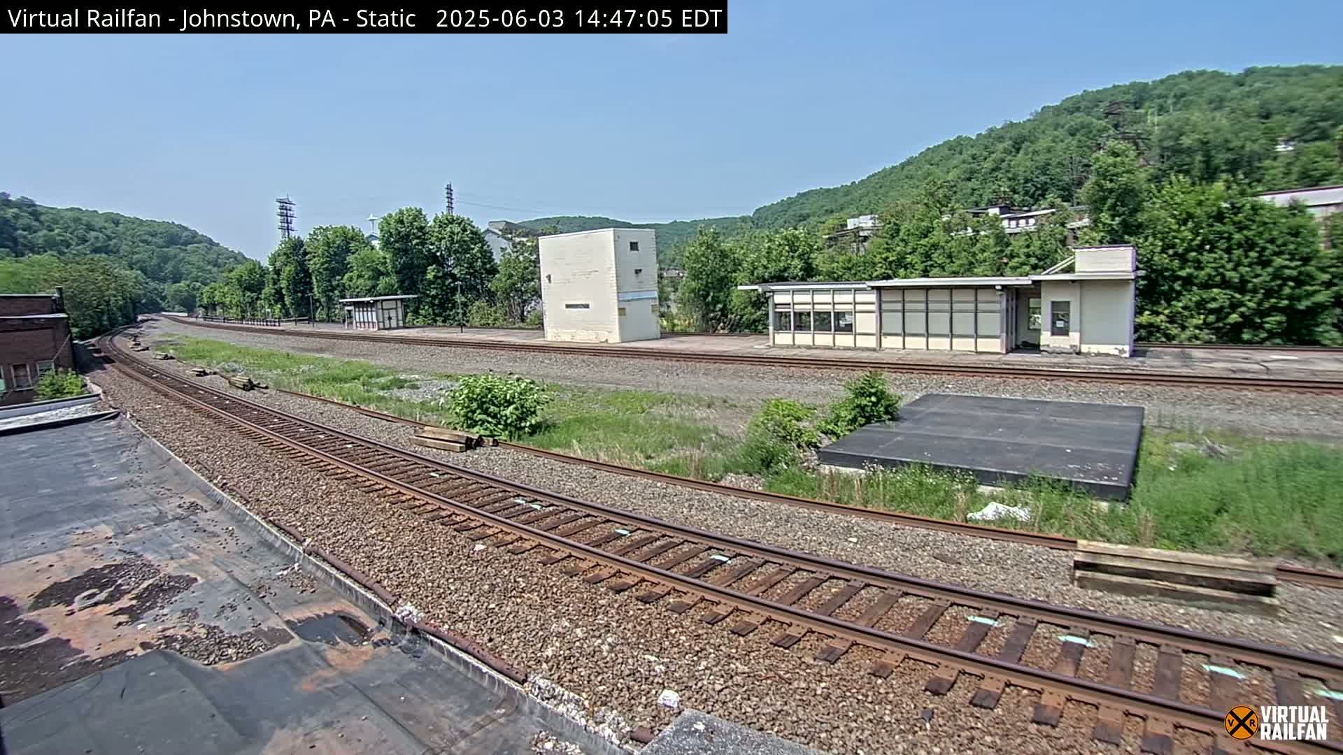 A sunny day reveals a railway track curving through a grassy area, past a small building and a taller, square structure, with green hills in the background.