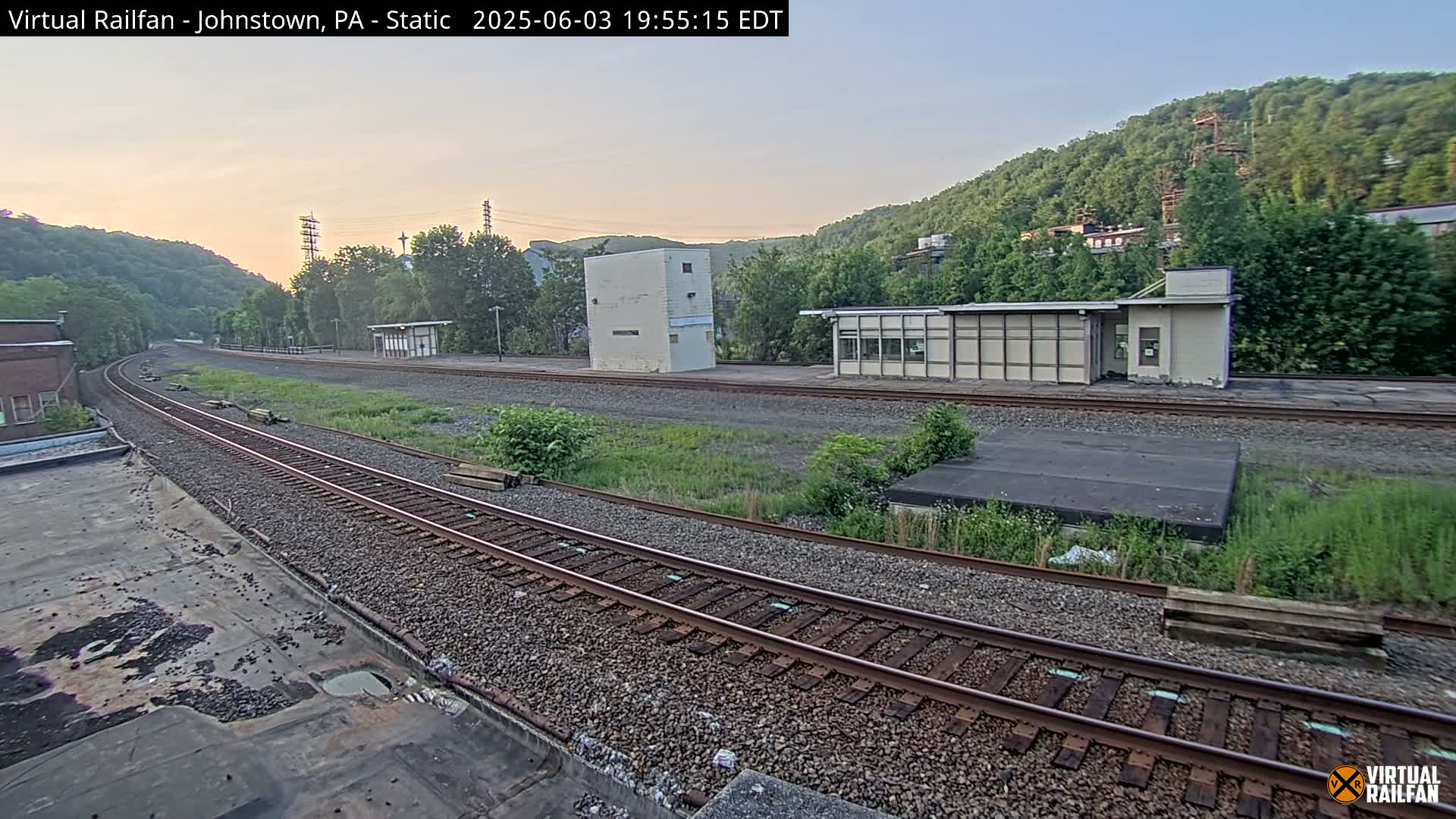 A train station with two small buildings sits beside a curving set of train tracks, all surrounded by green hills under a clear, twilight sky.
