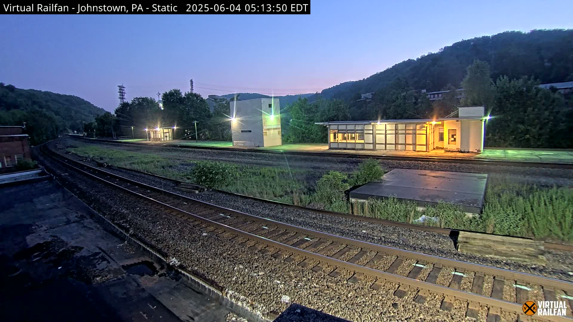 A train station with several buildings sits beside a curving set of train tracks at twilight under a clear sky.