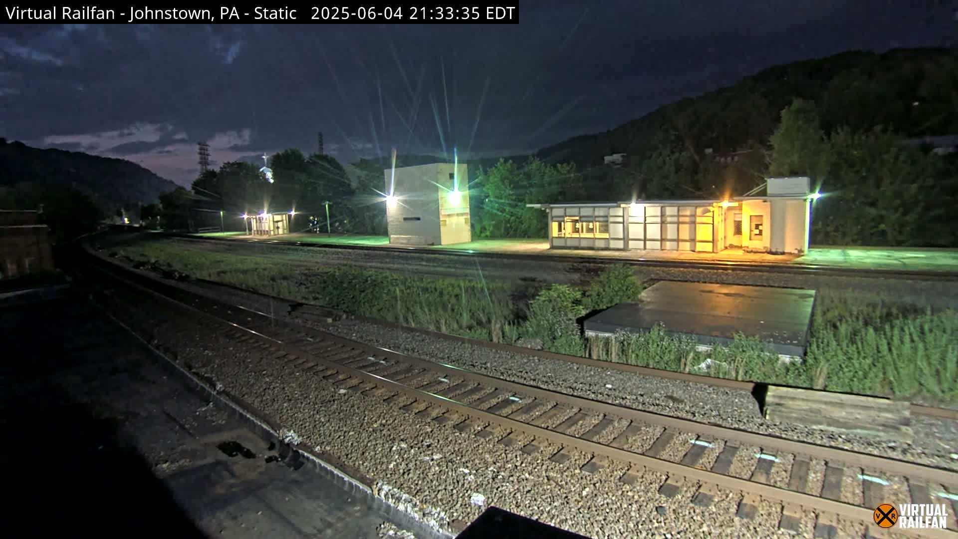 A nighttime view of a train station with several structures and railroad tracks, under a dark sky.