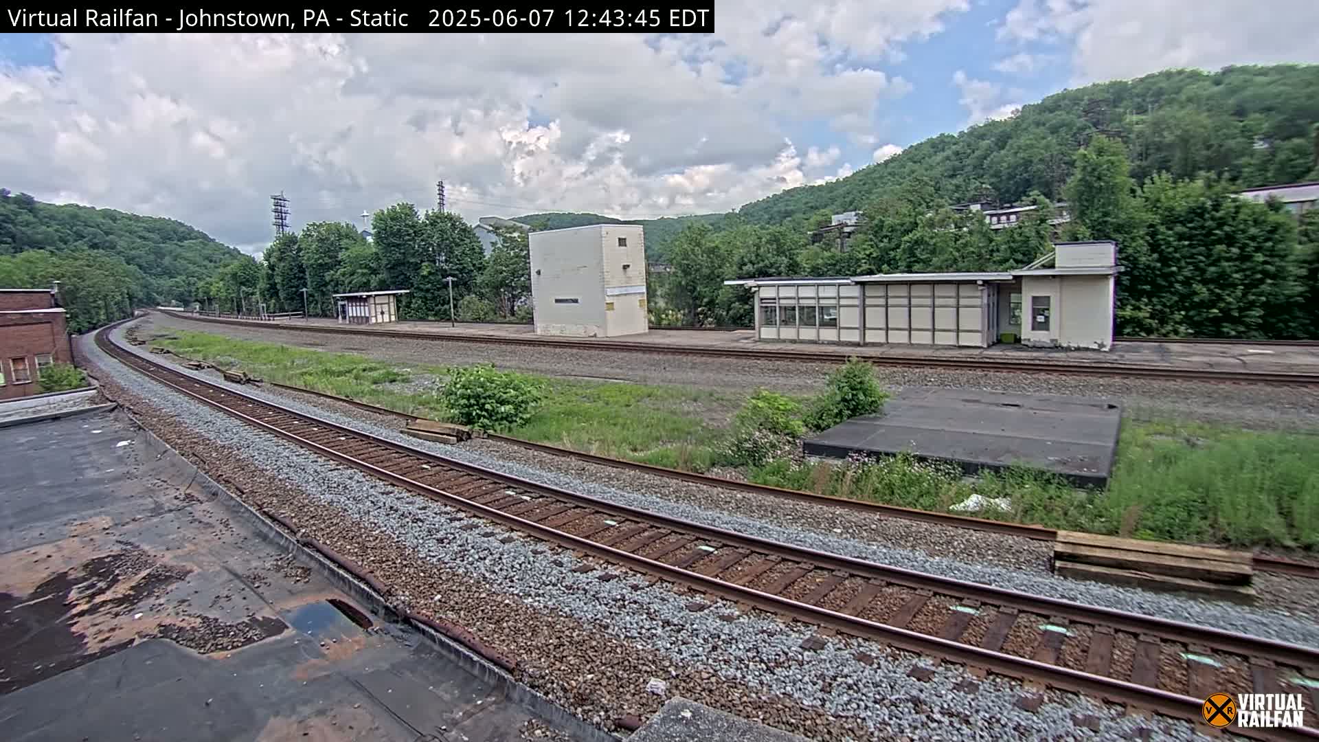 A train track curves gently through a railway yard on a partly cloudy day, with small buildings and overgrown vegetation flanking the tracks, nestled against a hillside covered in green trees.