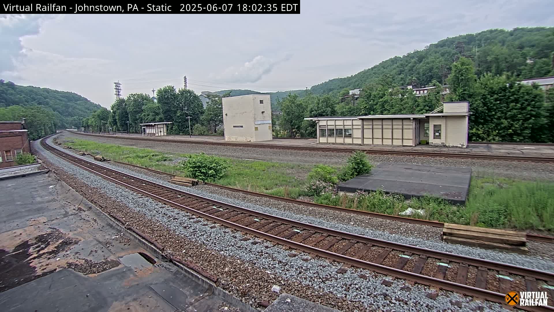 A mostly cloudy day shows a view of train tracks curving around a small station with several buildings, set against a backdrop of lush green hills.