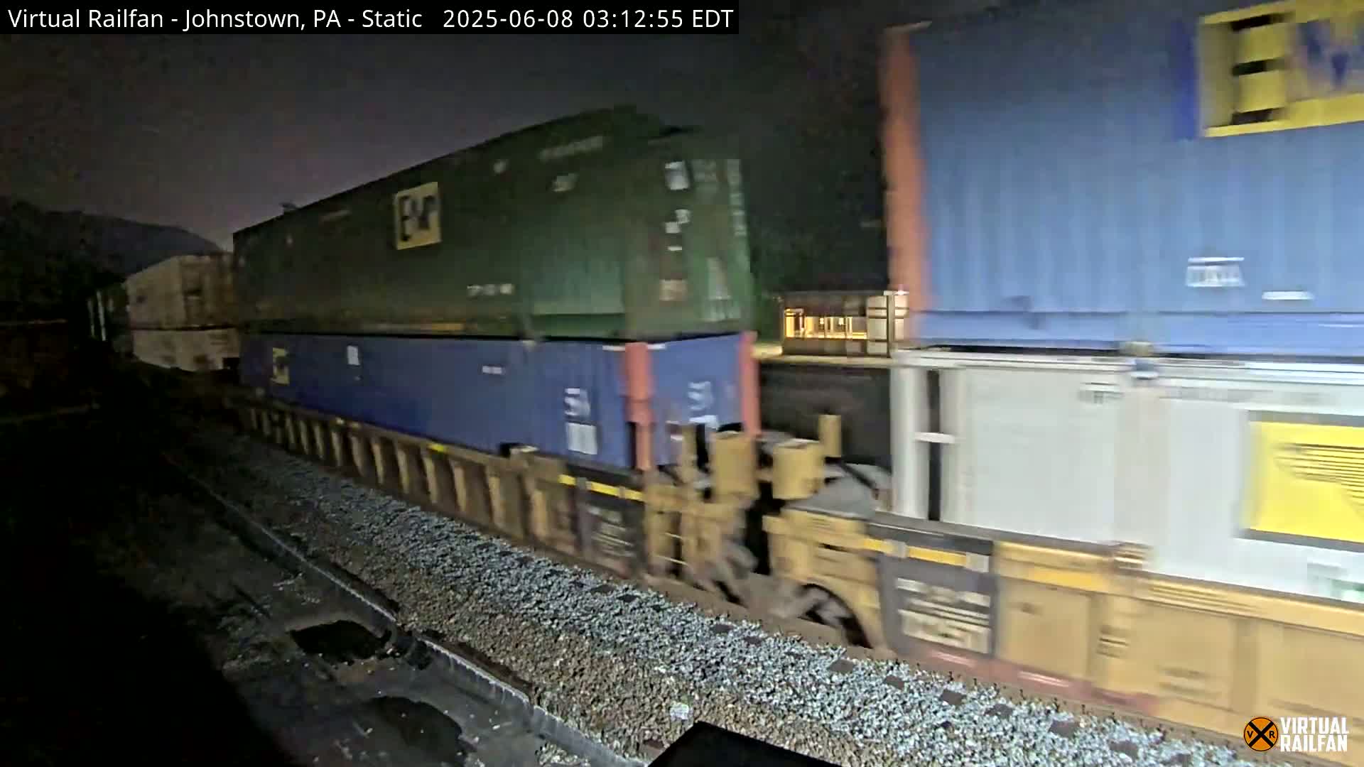 A freight train carrying various shipping containers moves along a track at night under a dark, overcast sky.