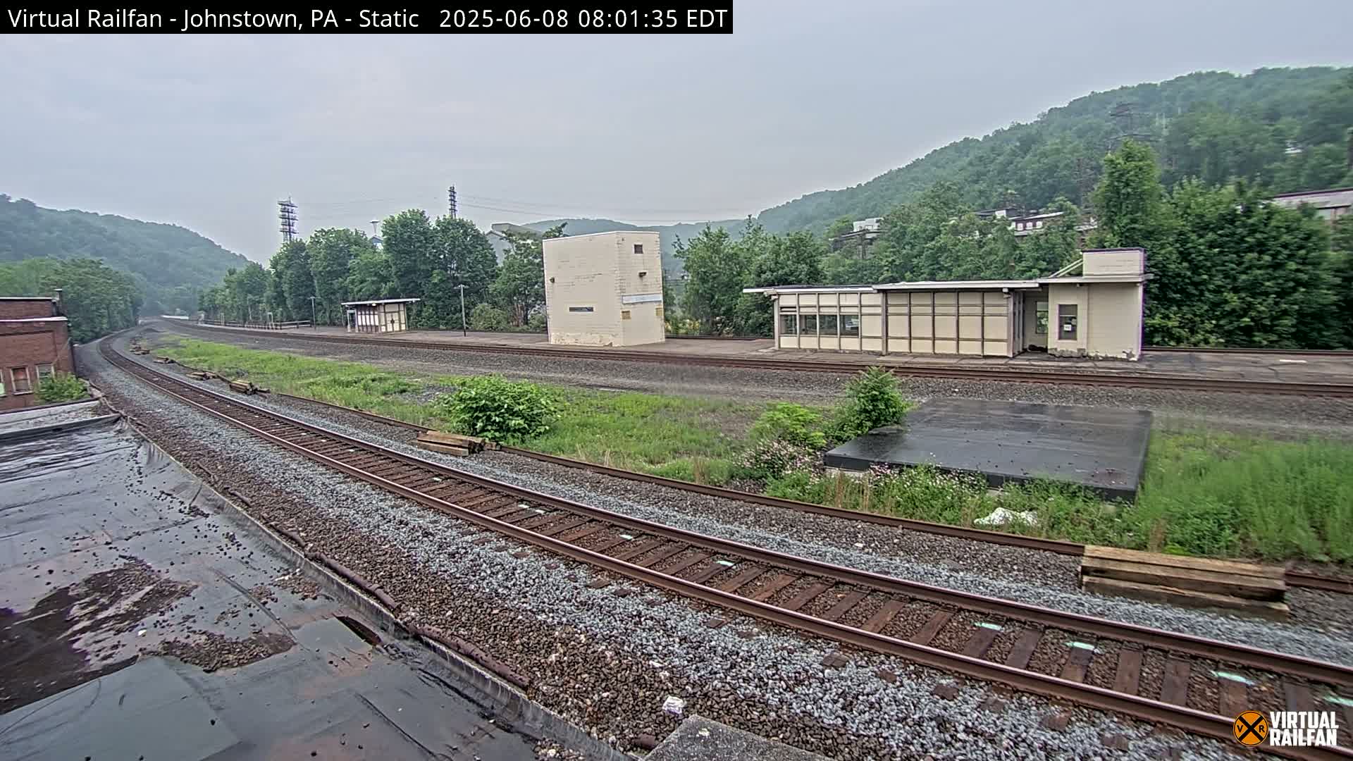 A view of a train station on an overcast day, showing multiple train tracks curving into the distance, bordered by grassy areas, small buildings, and a hillside covered in lush green trees.