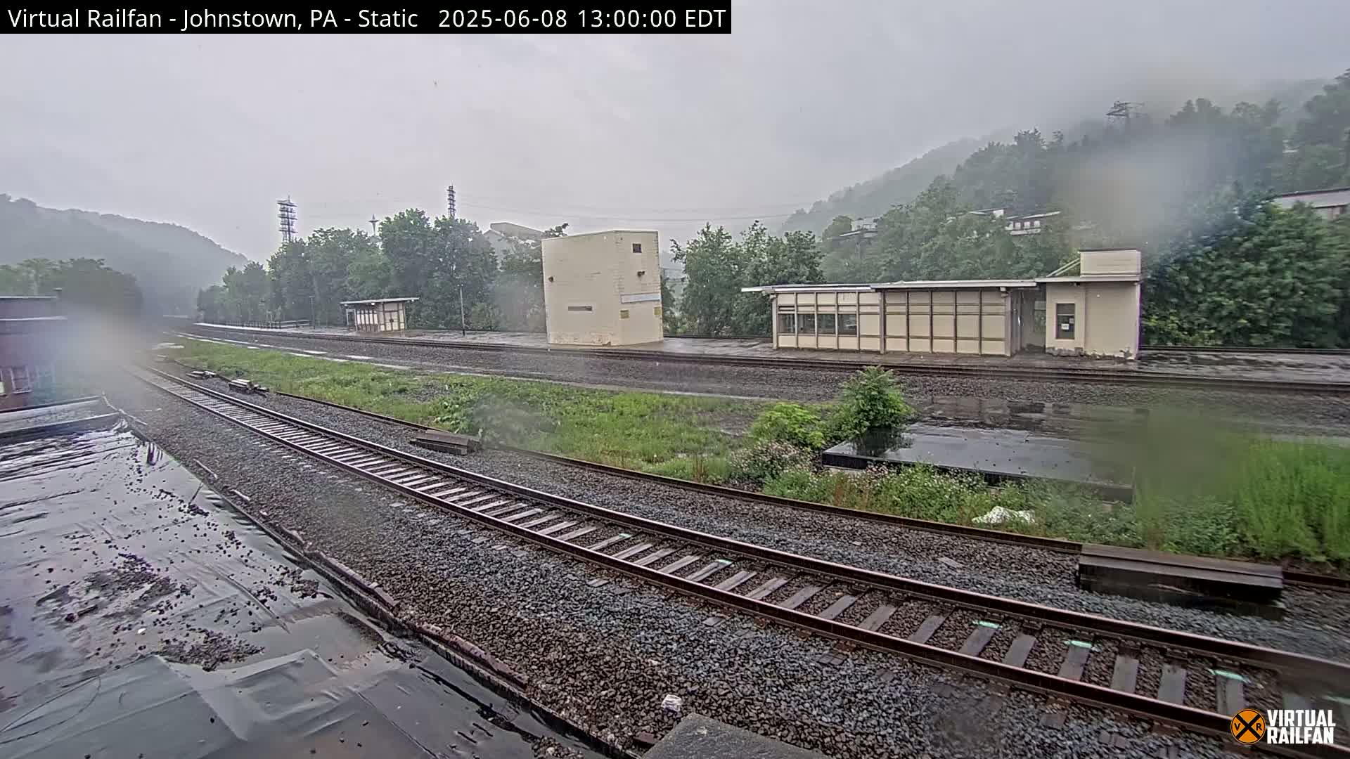 A rainy view of a railroad yard with multiple tracks, small buildings, and hills in the background.