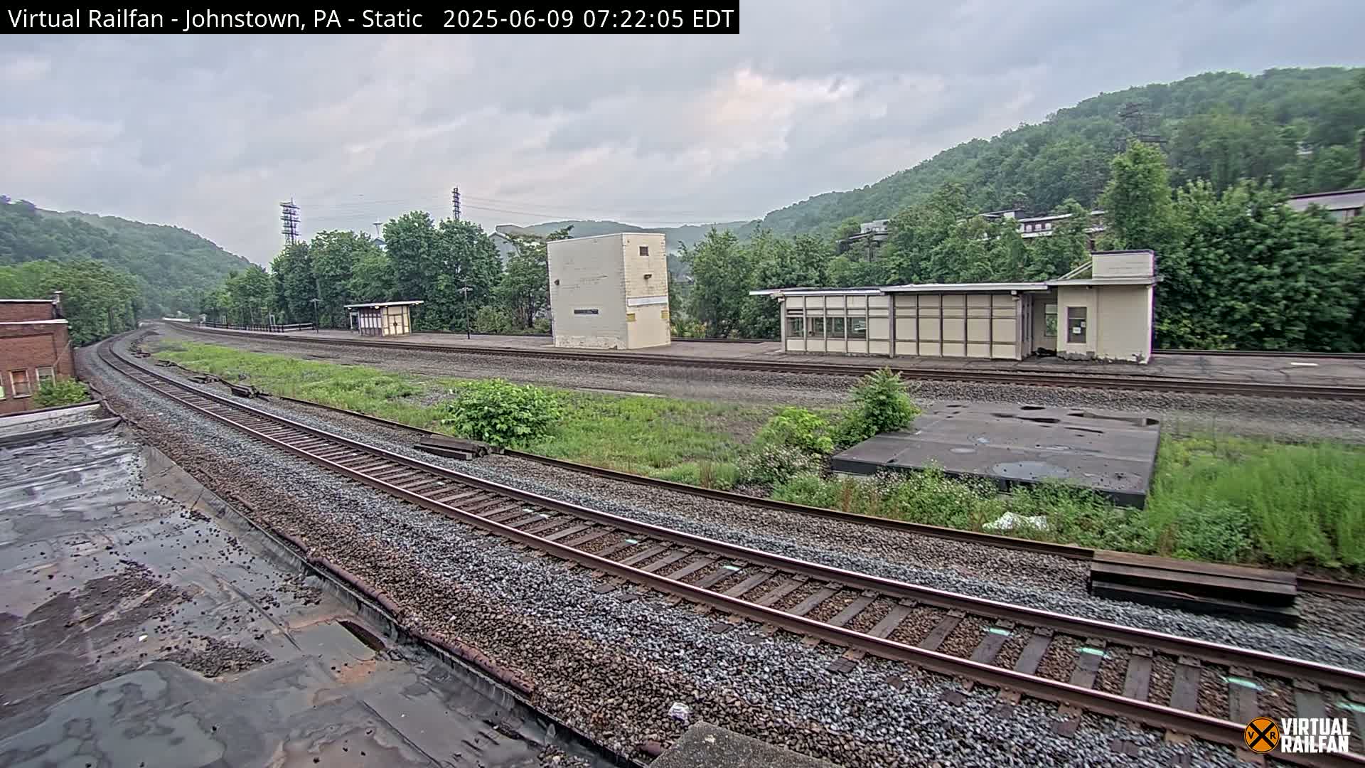 A curving train track stretches through a green, hilly landscape under an overcast sky, passing by several low buildings and overgrown vegetation.