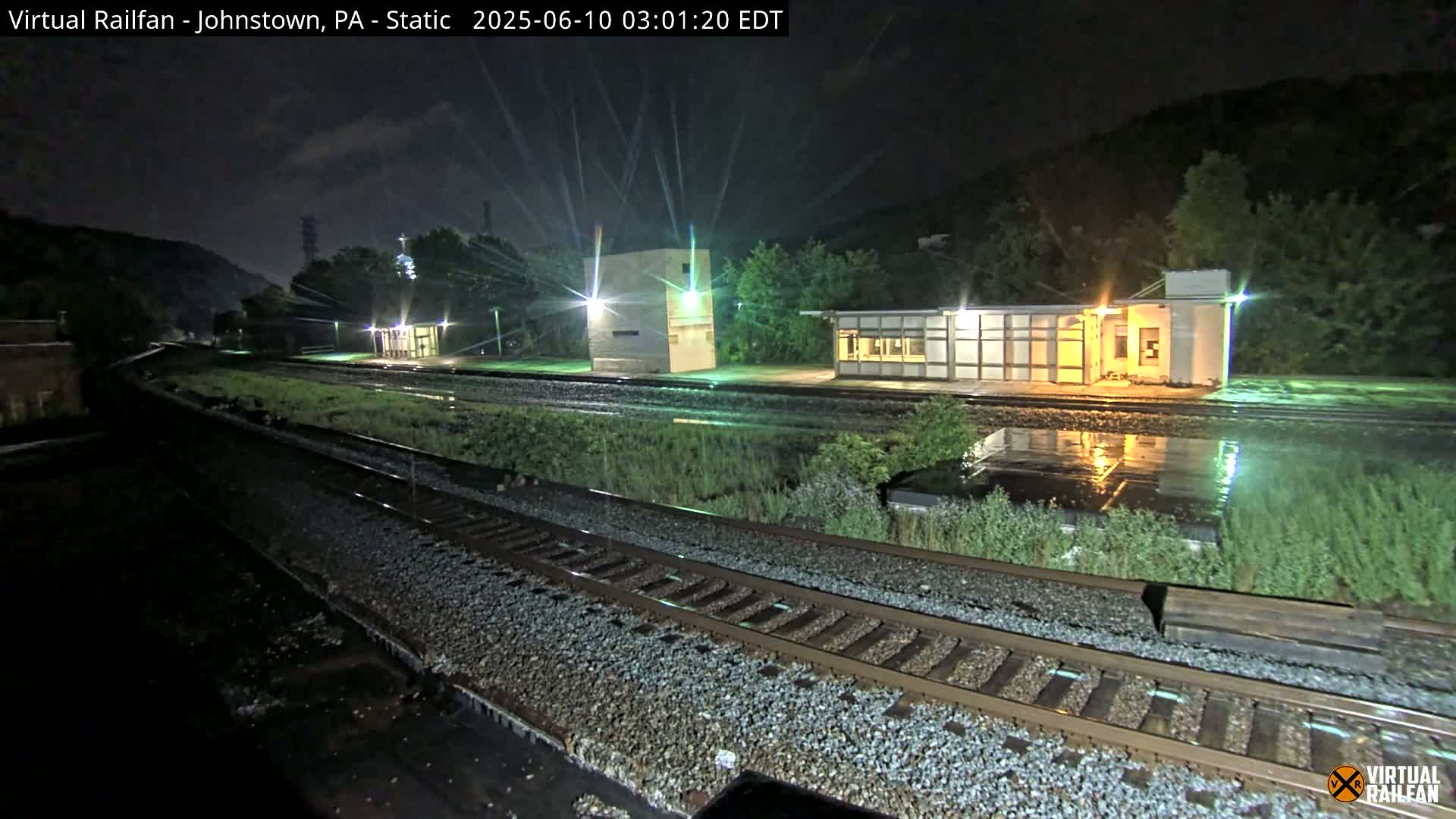 A nighttime view of a train station with two small buildings beside a set of train tracks, under wet conditions.