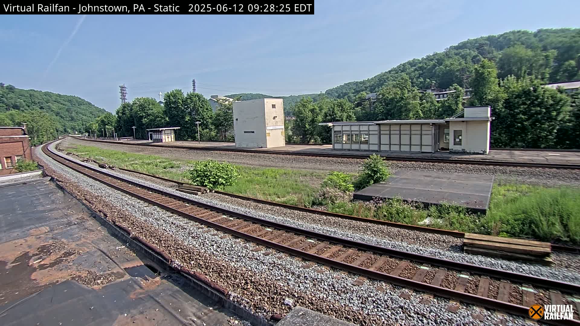A curving railroad track stretches into the distance beneath a clear blue sky, past several small buildings and a hillside covered in lush green vegetation.