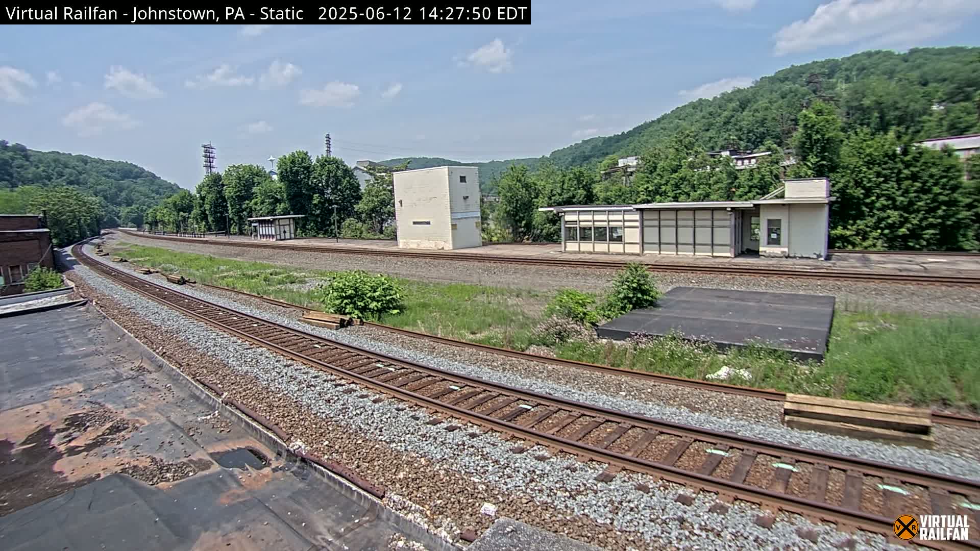 A train track curves through a grassy area with small buildings and a hillside covered in lush green trees under a partly cloudy sky.
