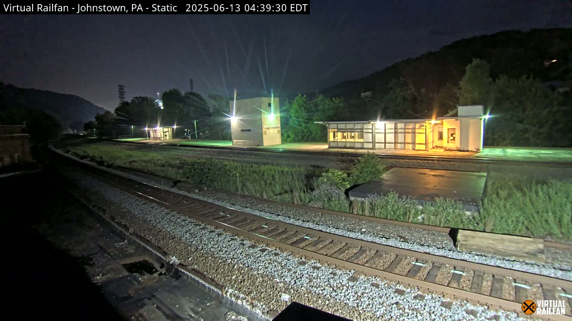 At night, a train station with several small buildings is illuminated, situated beside a set of train tracks that extend into the darkness against a backdrop of hills.
