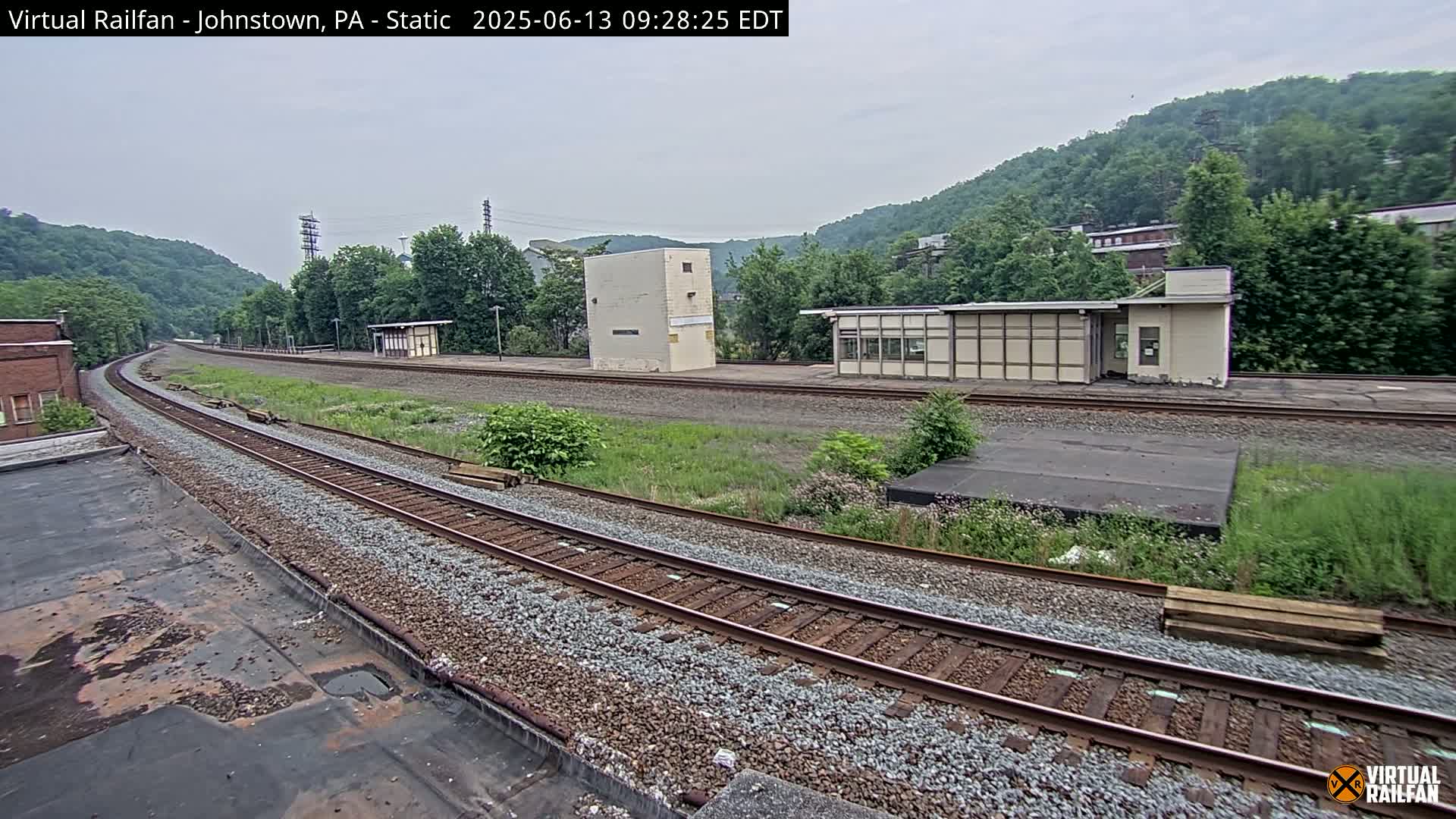 A view of railroad tracks curving away from the camera toward a hillside under an overcast sky, with several small buildings visible along the tracks.