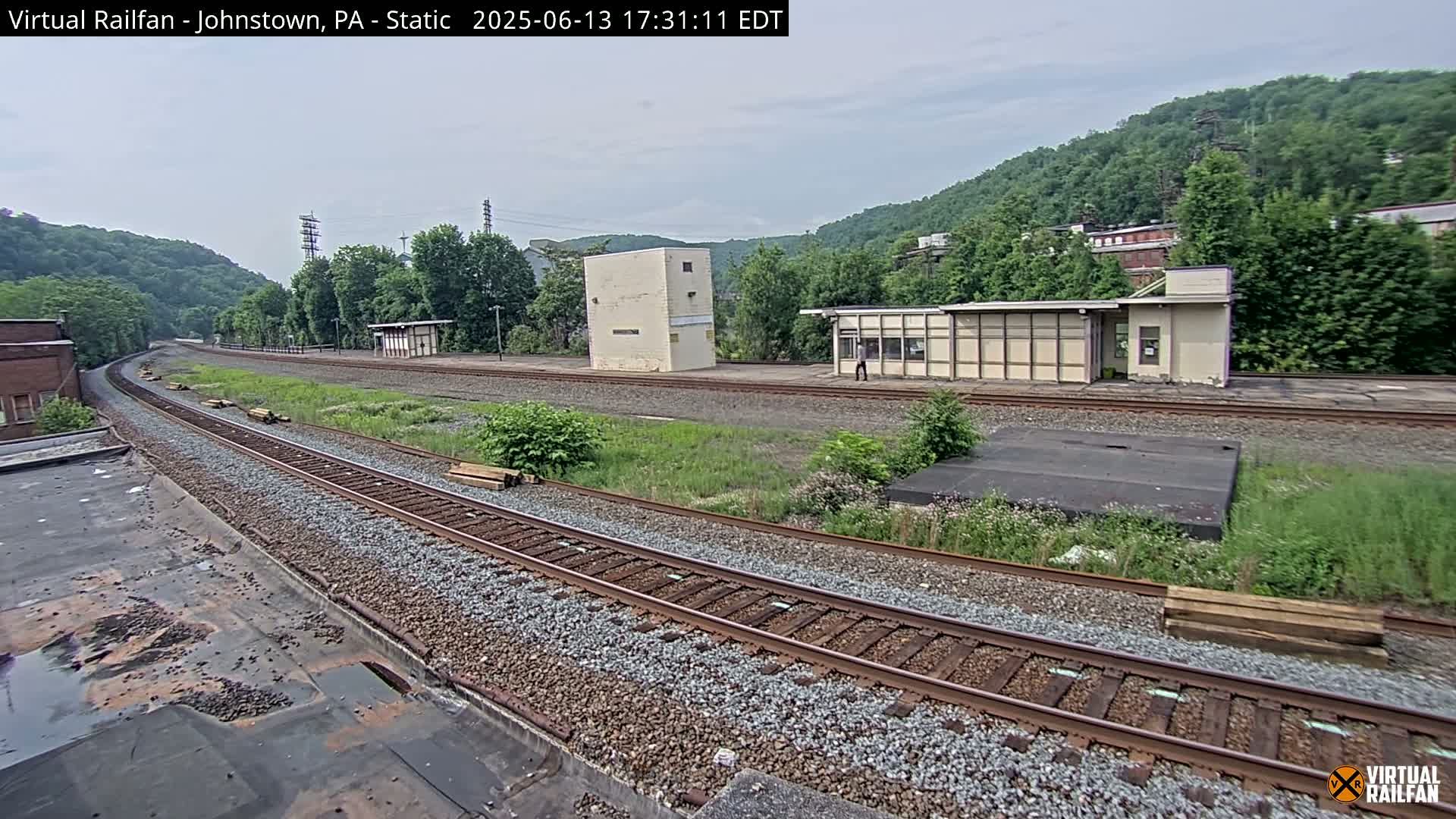 A view of railroad tracks curving through a grassy area with several small buildings and a hill covered in lush green trees under an overcast sky.