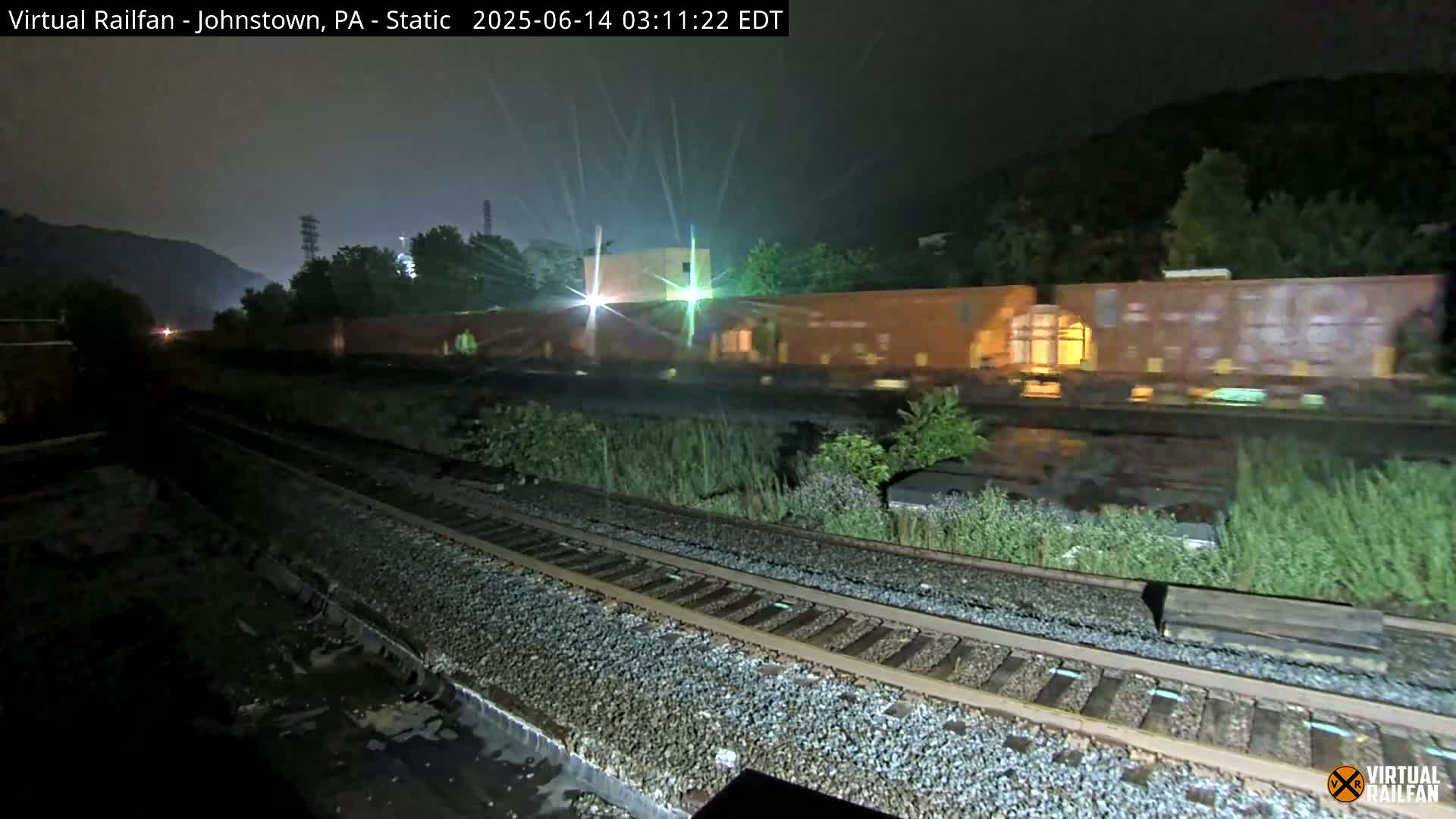 A freight train passes by at night in the rain, next to railroad tracks bordered by vegetation and hills in the background.