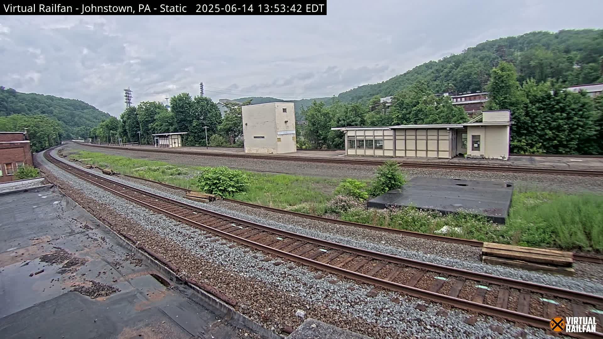 A curving railway track extends into the distance, passing by several small buildings and overgrown vegetation under an overcast sky.