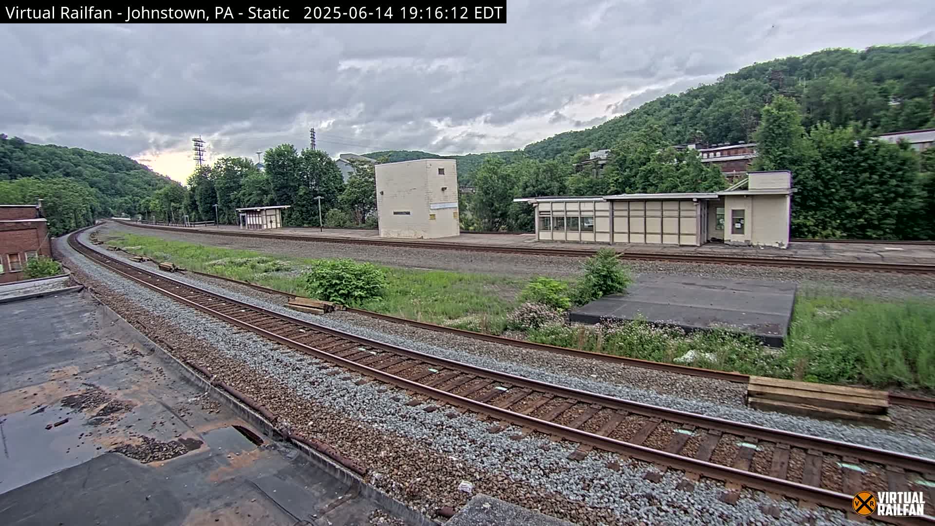 A curving railway track extends into the distance, passing by several small buildings and overgrown vegetation under an overcast sky.