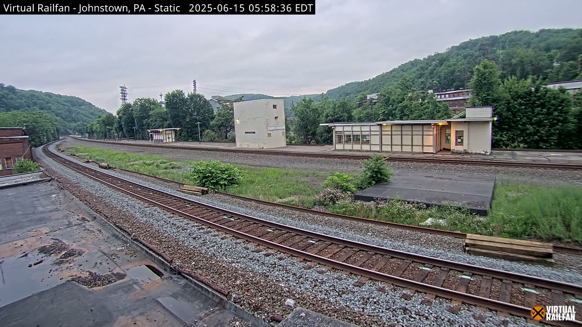 A view of a train track curving through a grassy area with two small buildings and hills in the background under an overcast sky.