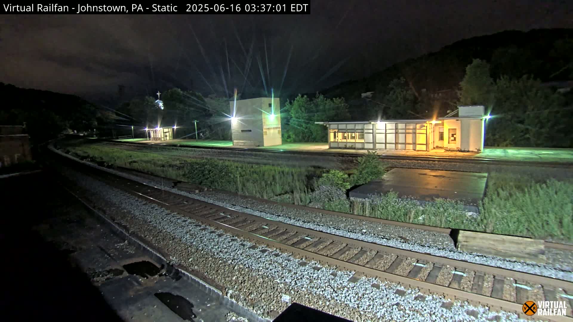 At night, a train track curves gently past a small, illuminated train station and other structures, all set against a dark hillside.