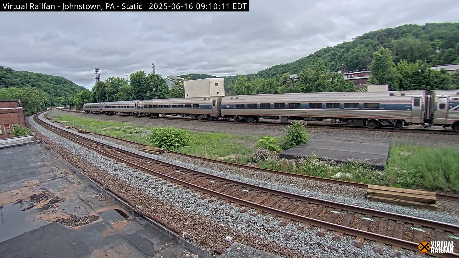 A long passenger train passes by on a set of train tracks under an overcast sky, with a hillside visible in the background.